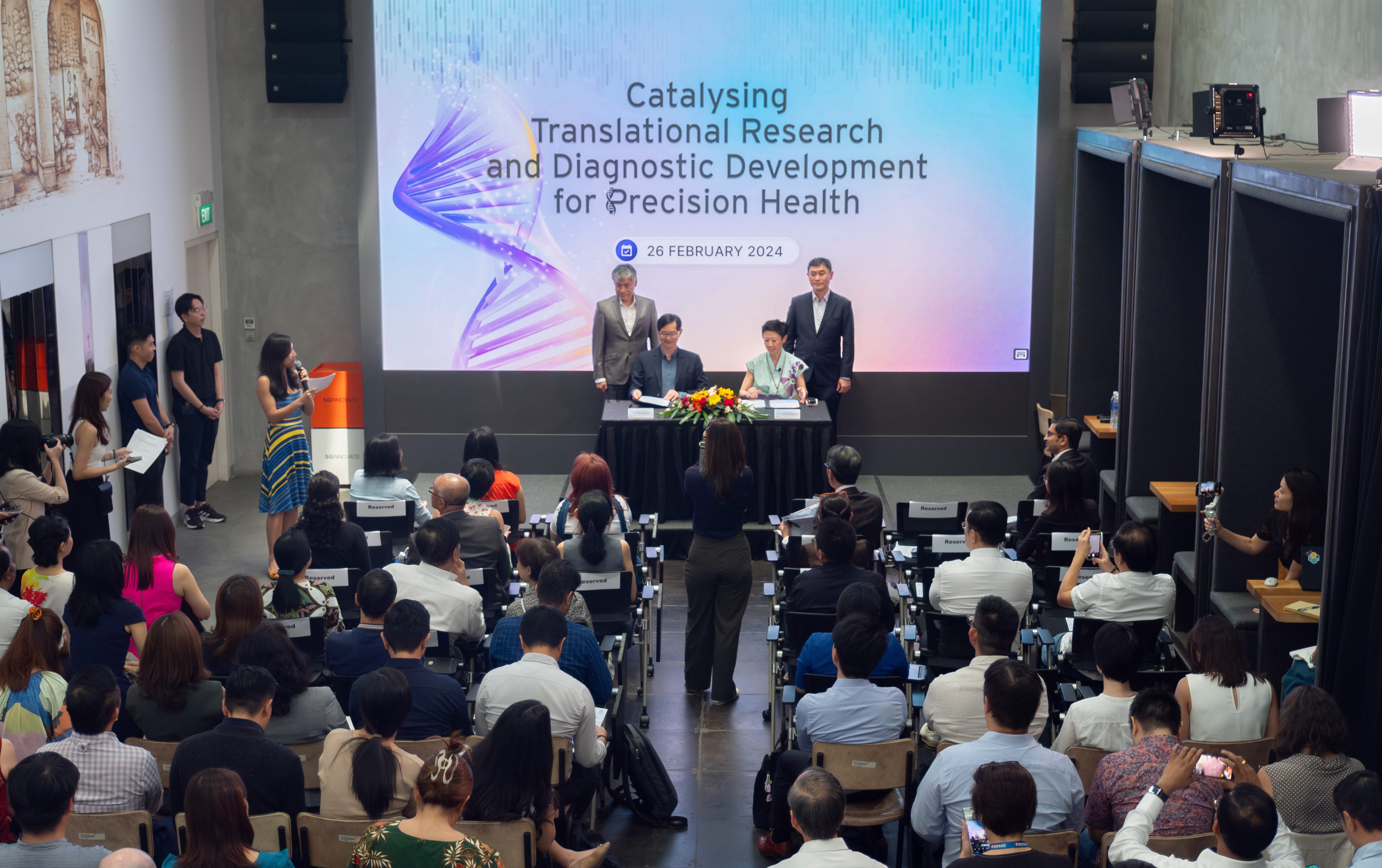 A large audience listens to a woman speak at a podium in front of a screen displaying a conference title on precision health.