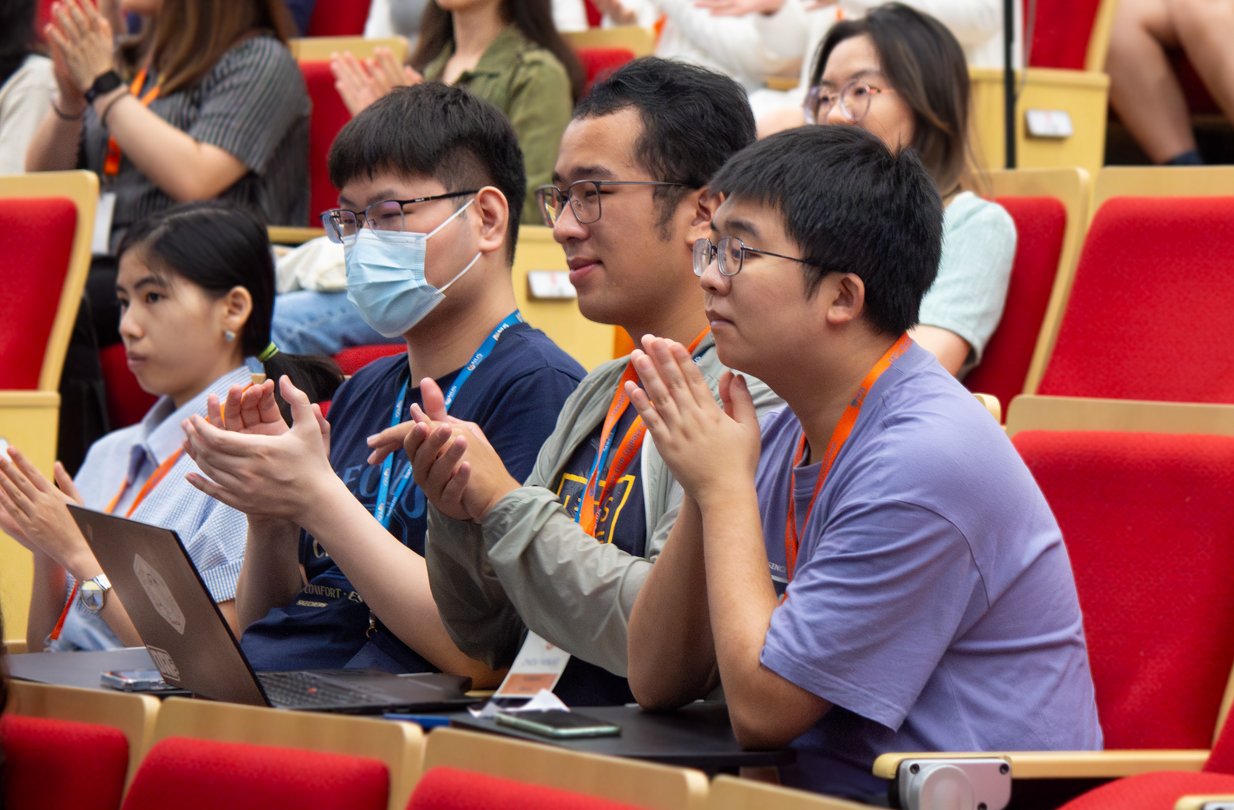 People sitting in a lecture hall and clapping with red seats, laptops on desks, and wearing conference badges.