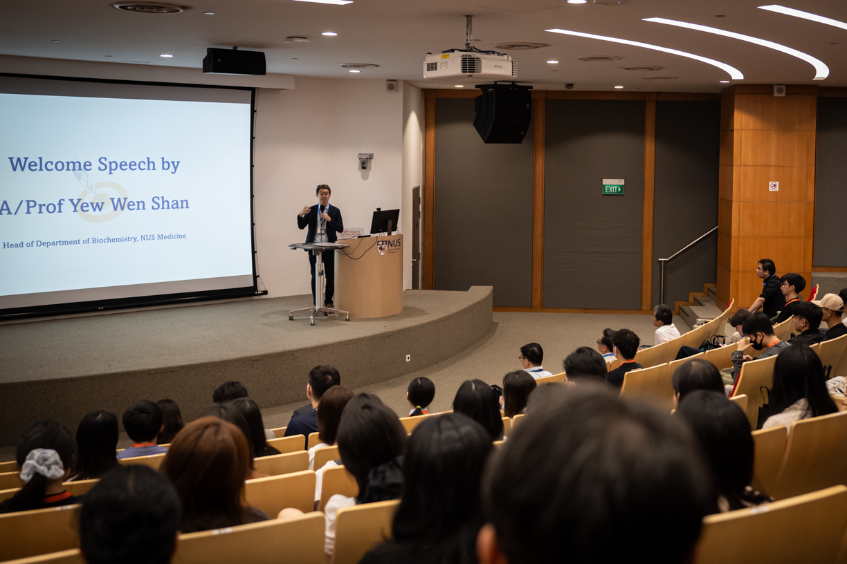 A speaker is giving a presentation in an auditorium full of people, with a slide displaying a welcome speech by A/Prof Yew Wen Shan.