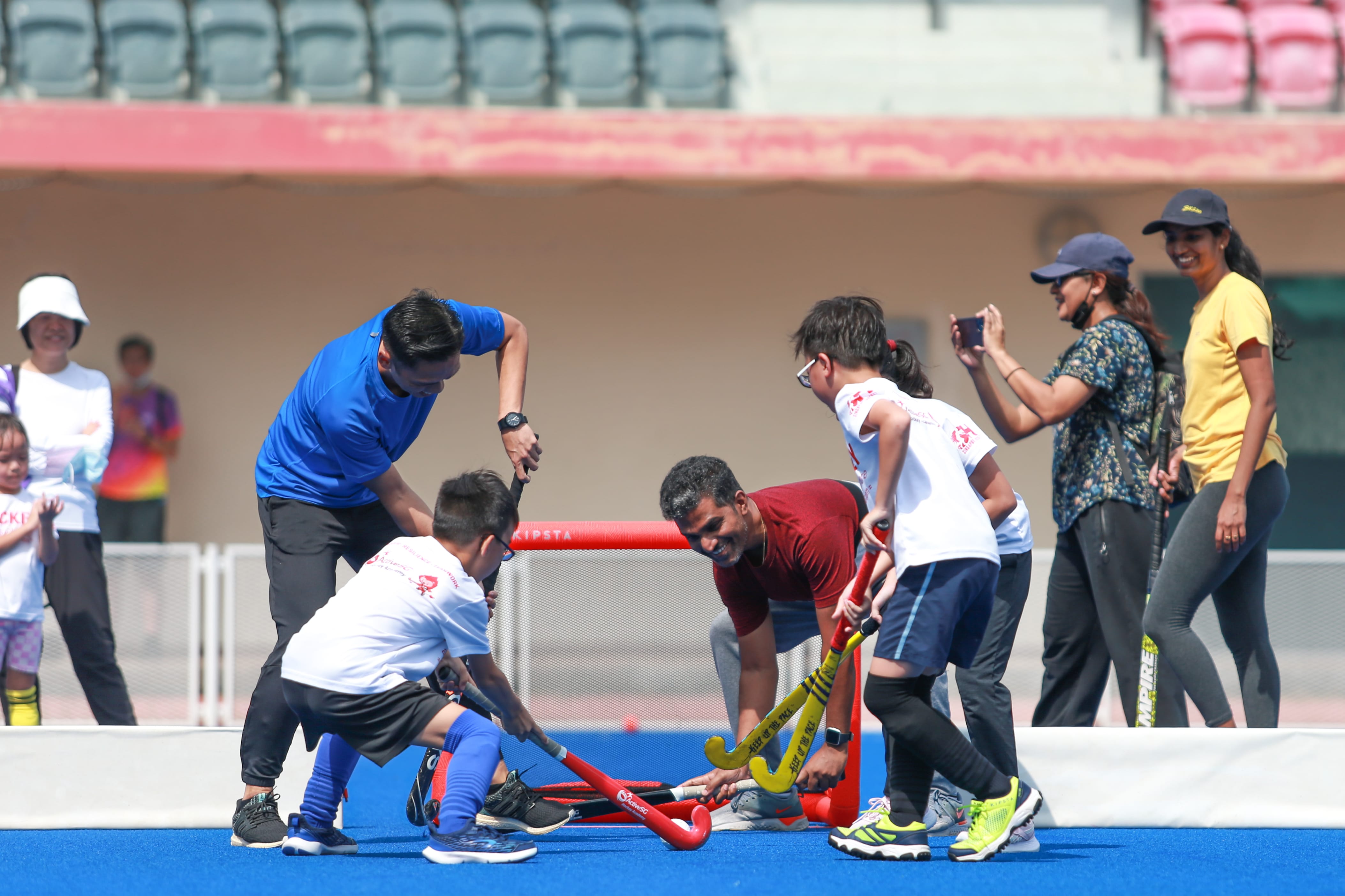 Children in white shirts and blue shorts play field hockey, guided by adults in a stadium.