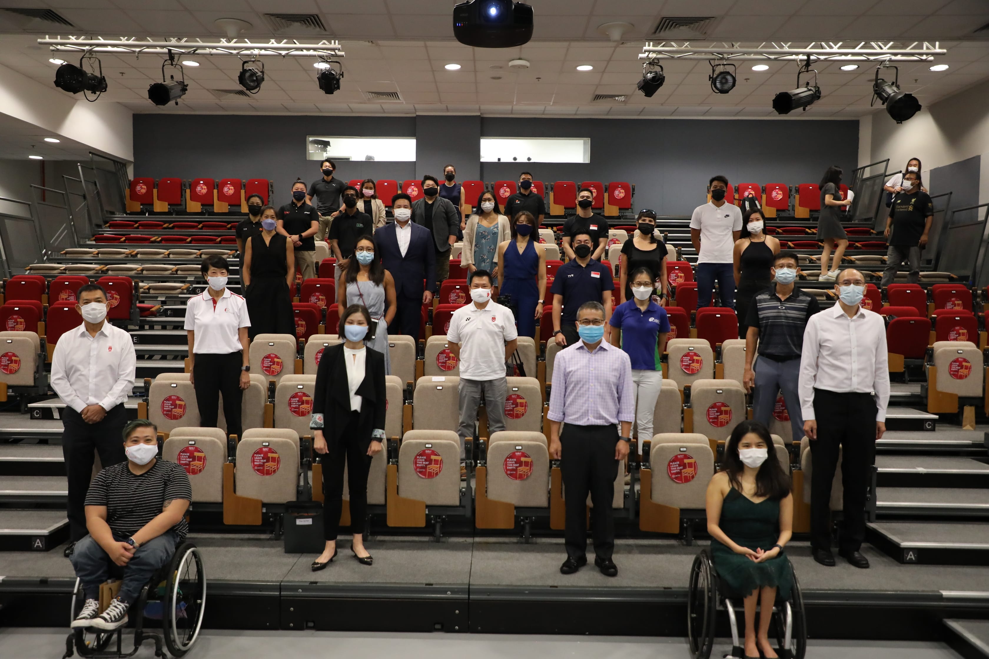 People wearing various outfits standing and sitting in a tiered auditorium with red and beige seats.