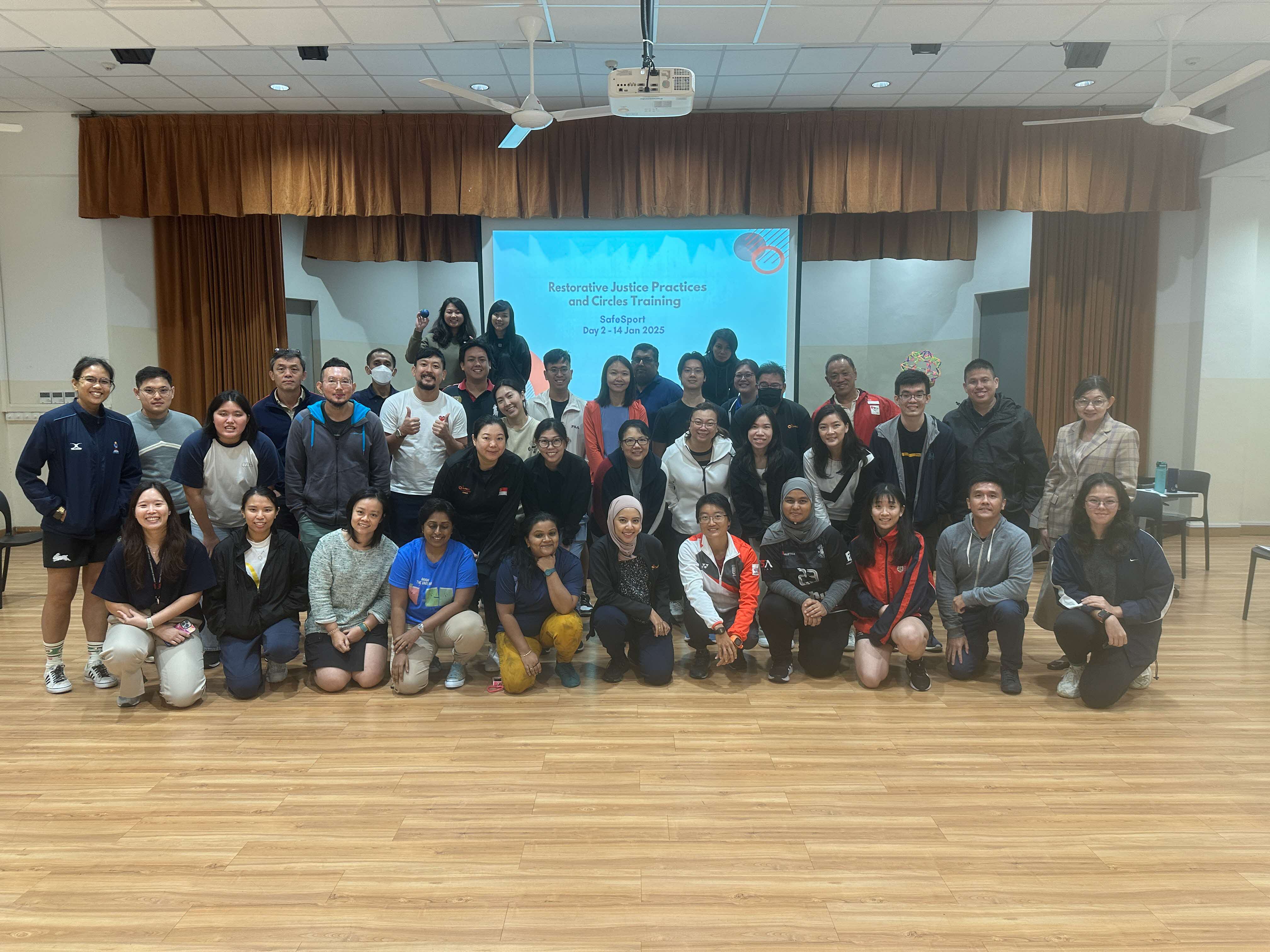 A large group of people posing indoors, in front of a presentation screen displaying "Restorative Justice Practices and Circles Training".