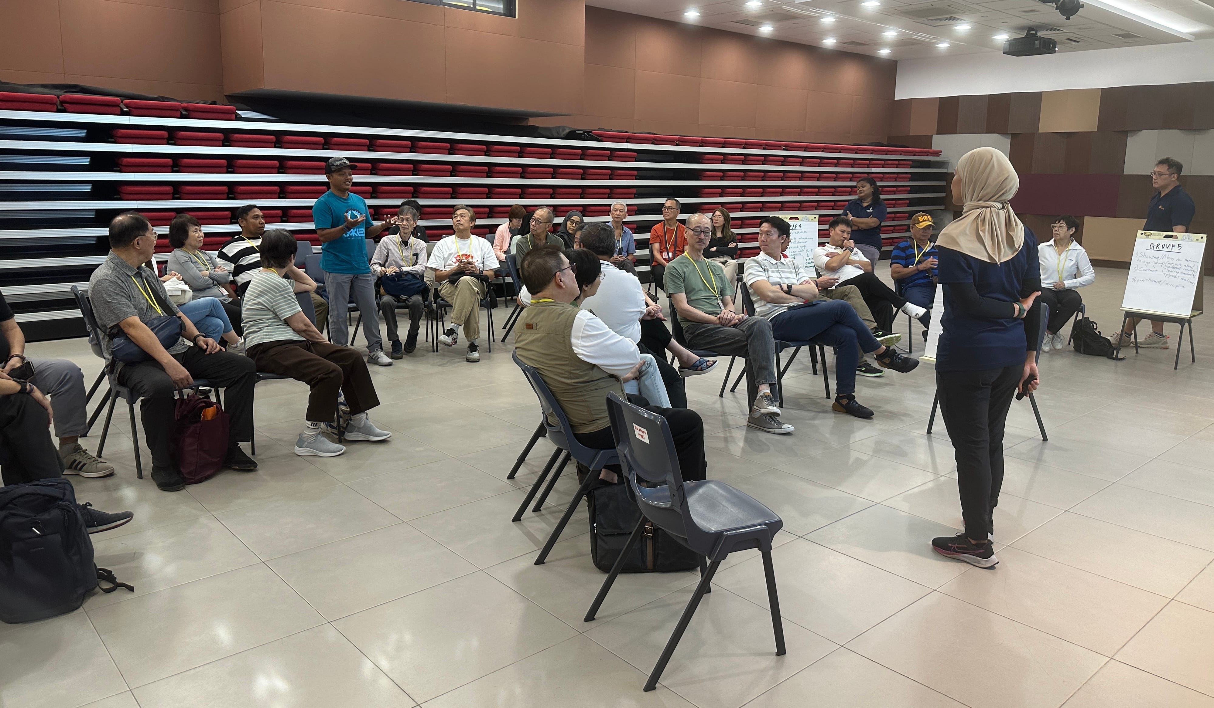 People sitting in a circle on chairs in a room, listening to a person speaking while another stands near a whiteboard.