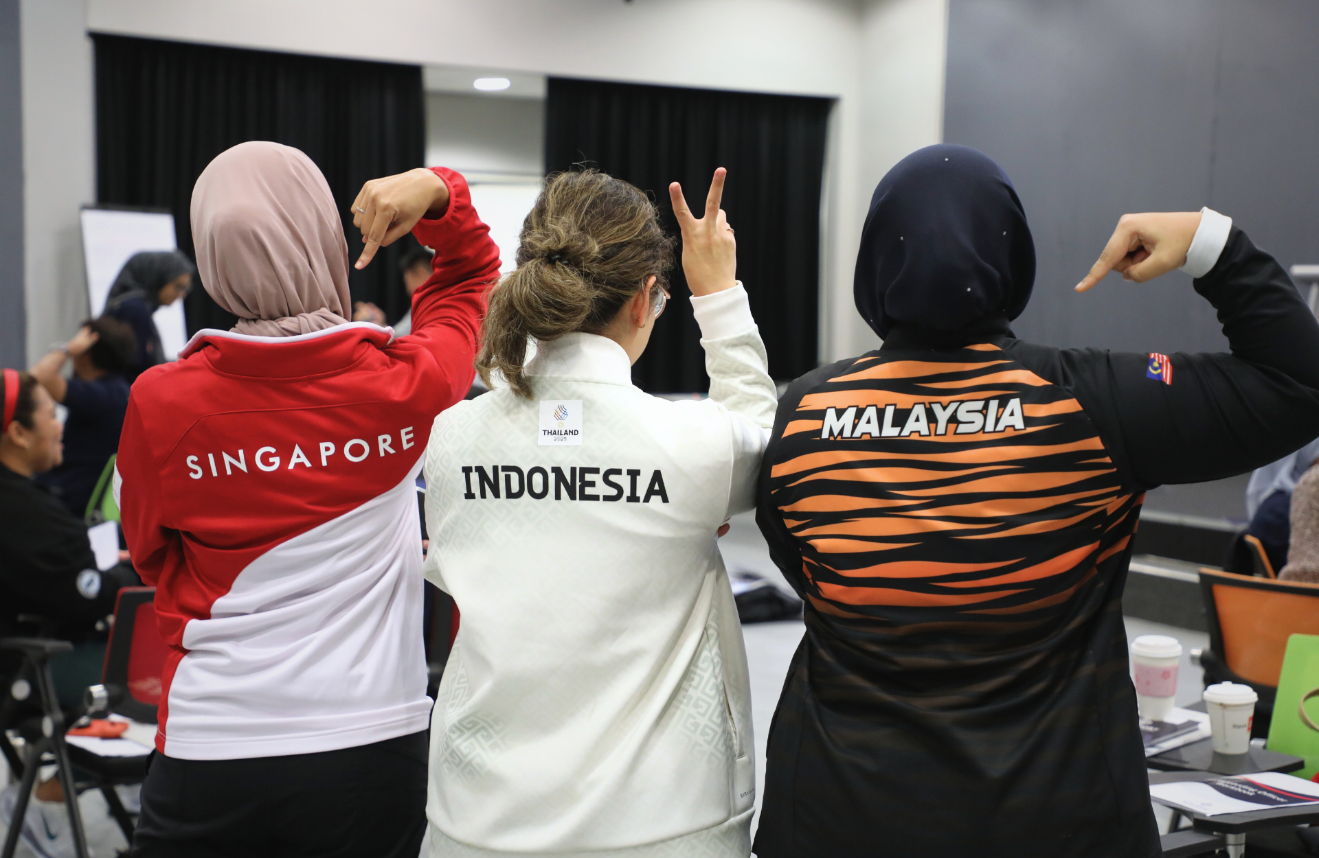 Three people stand with their backs to the camera, displaying jackets labeled Singapore, Malaysia and Indonesia. They point at each other signifying togetherness.