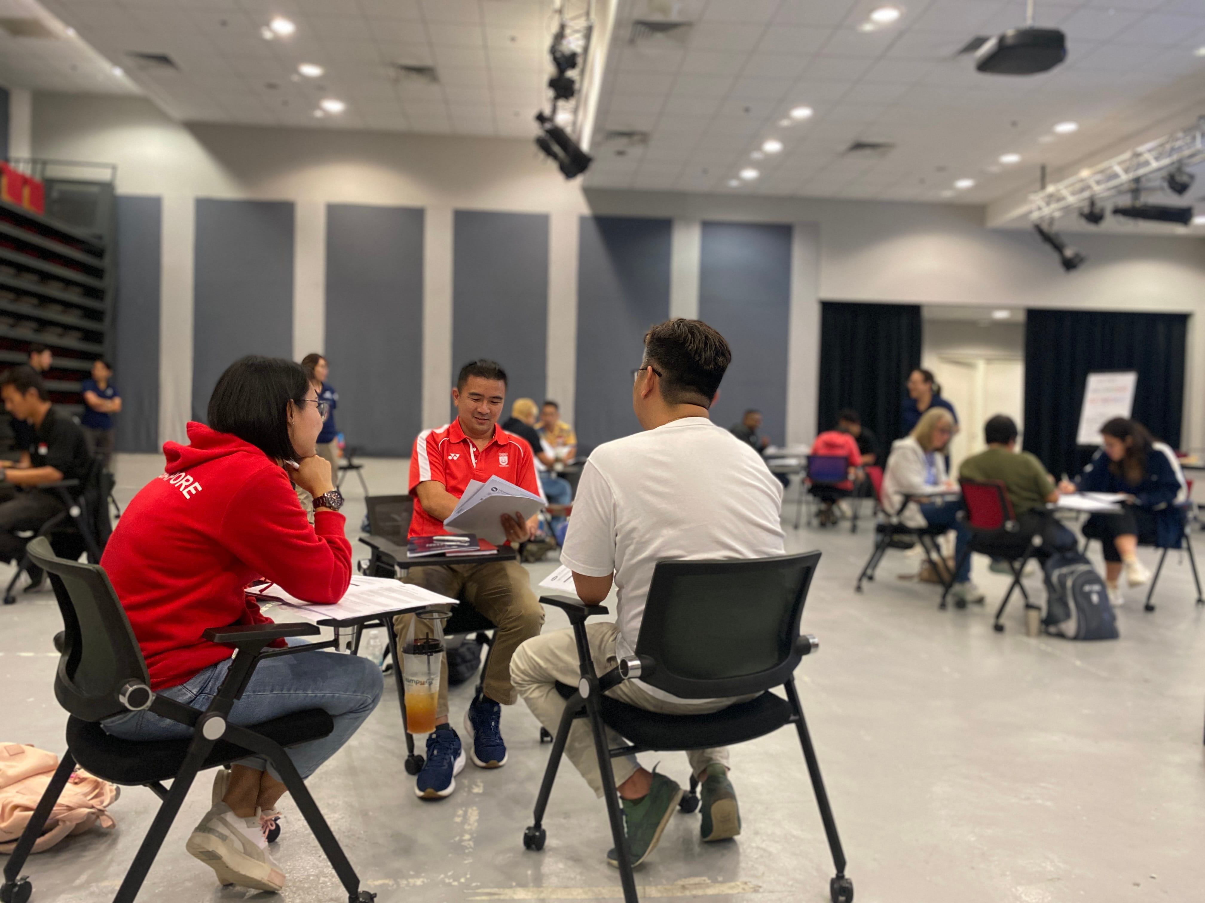 A group of people in a large room sit around tables, facing each other, engaging in discussions.