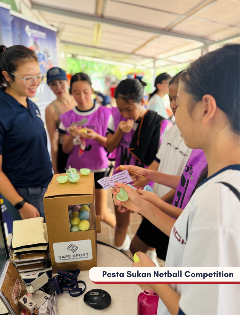 People are gathered around a table with a cardboard capsule machine, filled with colourful capsules, at Pesta Sukan Netball Competition.