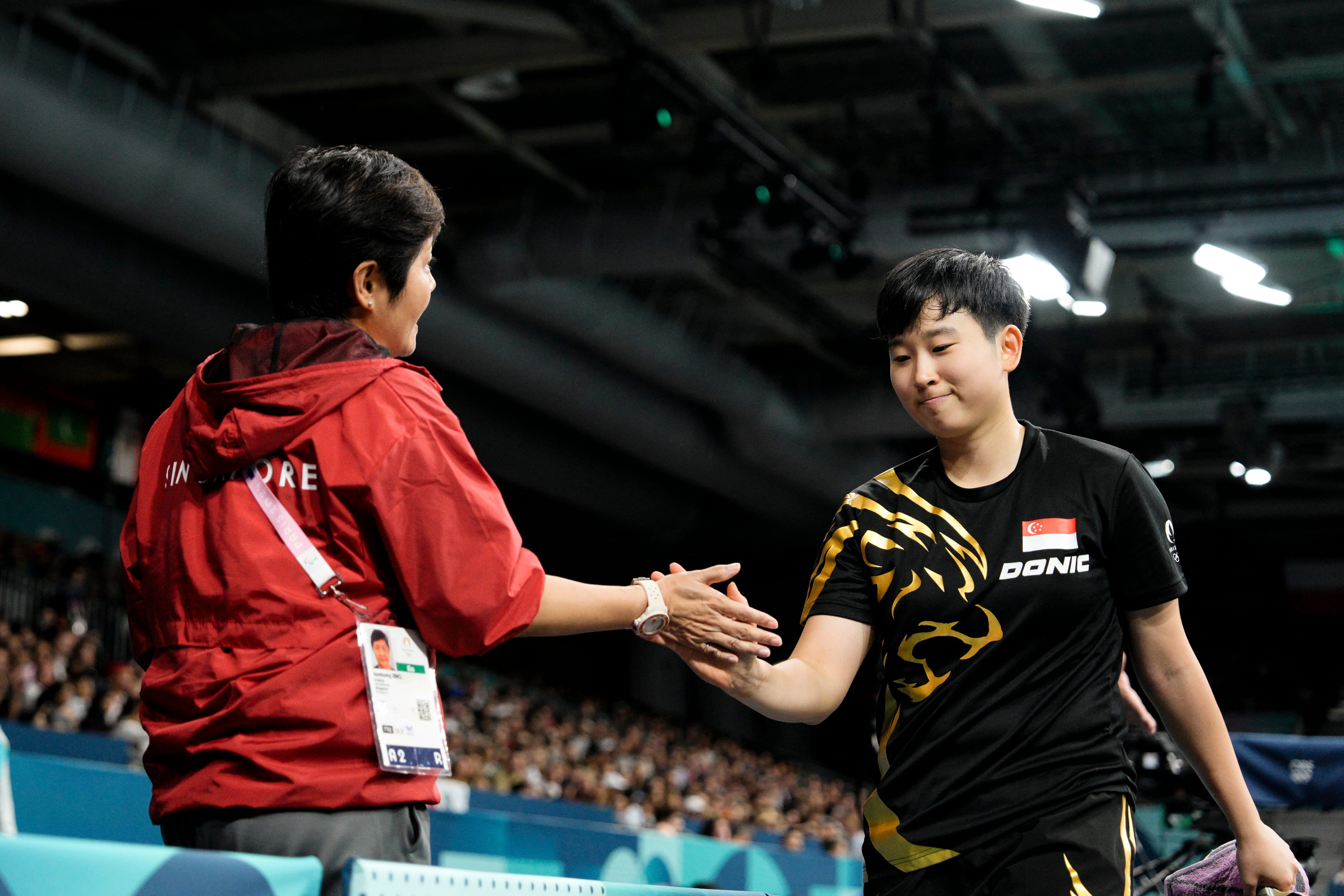 Two people shake hands; one person wears a red jacket with 'Singapore,' and the other a black shirt with a tiger design.