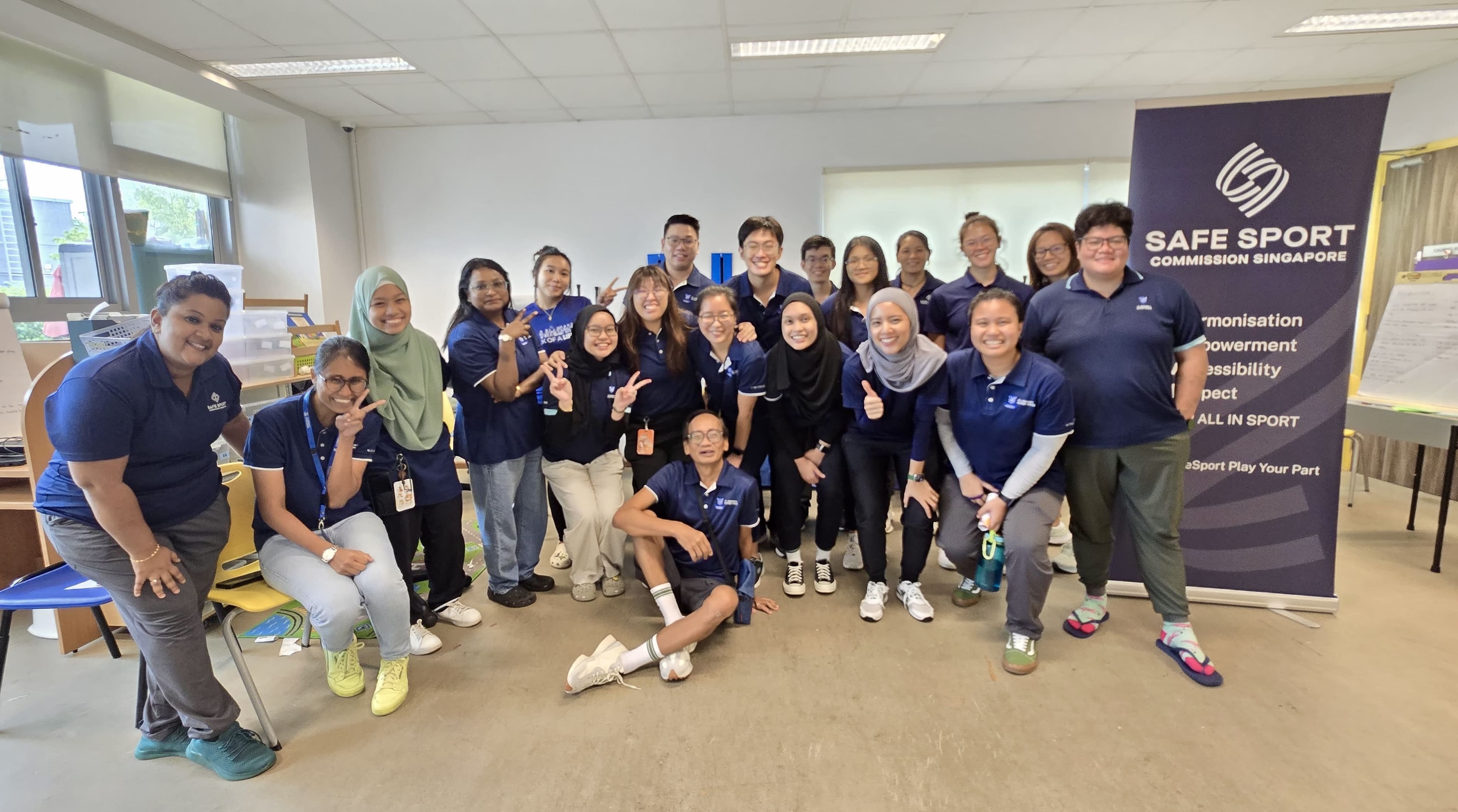 A group of people wearing navy blue shirts are posing together indoors, beside a "Safe Sport Commission Singapore" banner.