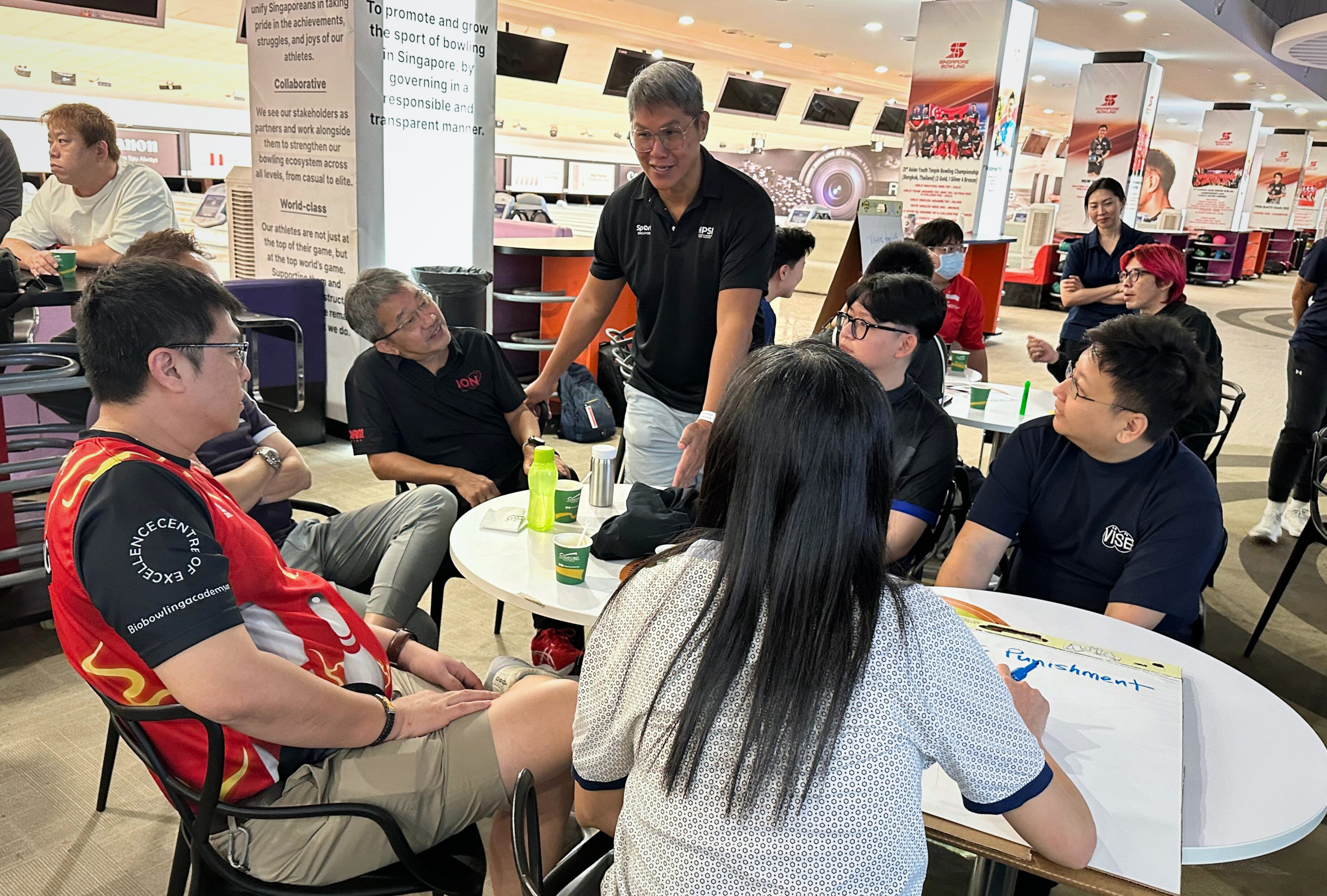 A group of people are sitting and standing around a table in a bowling center, talking and drinking beverages.