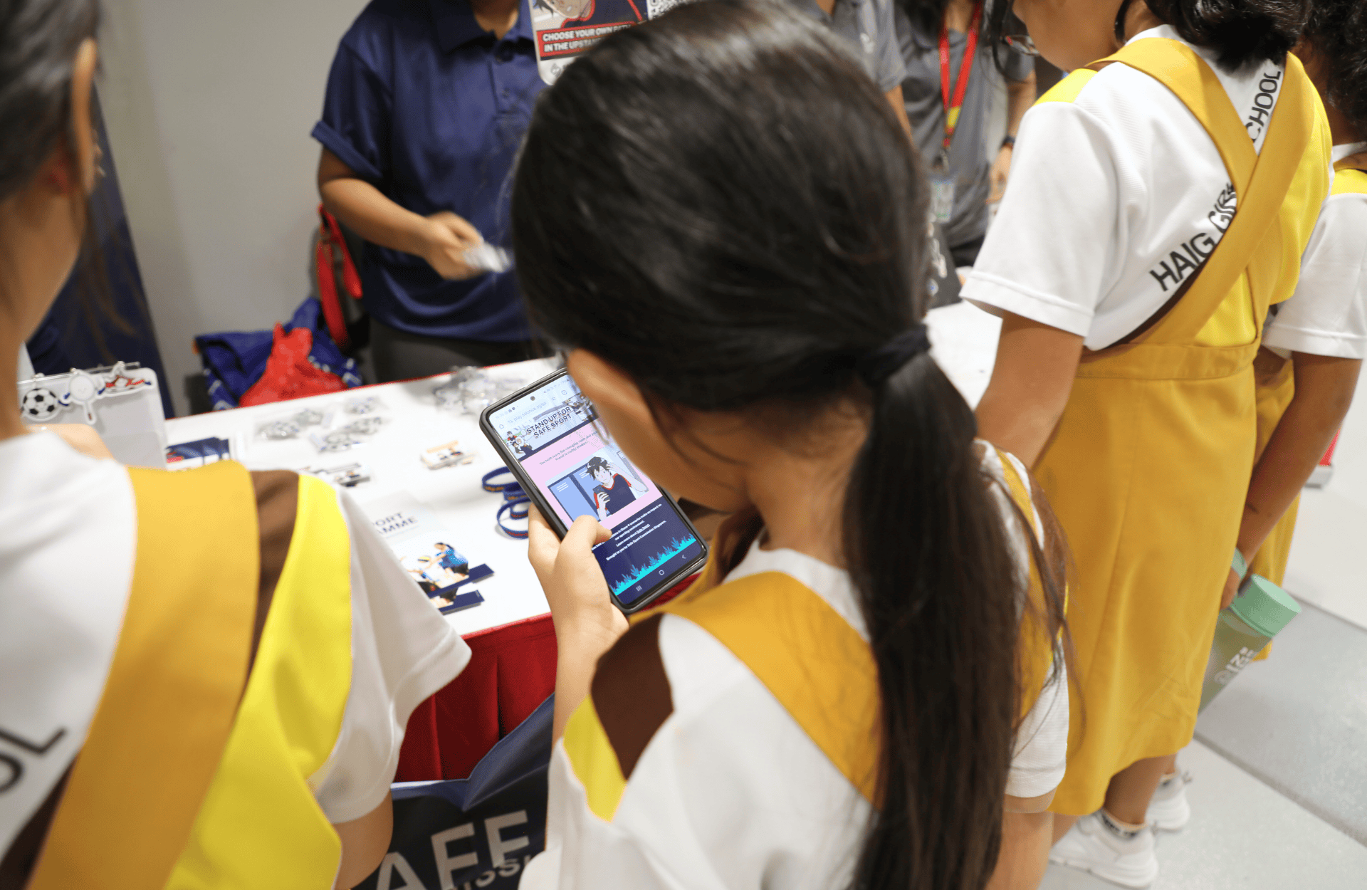 A young student in a yellow uniform looks at a smartphone near a booth. Others in similar attire stand around, conveying curiosity and engagement.