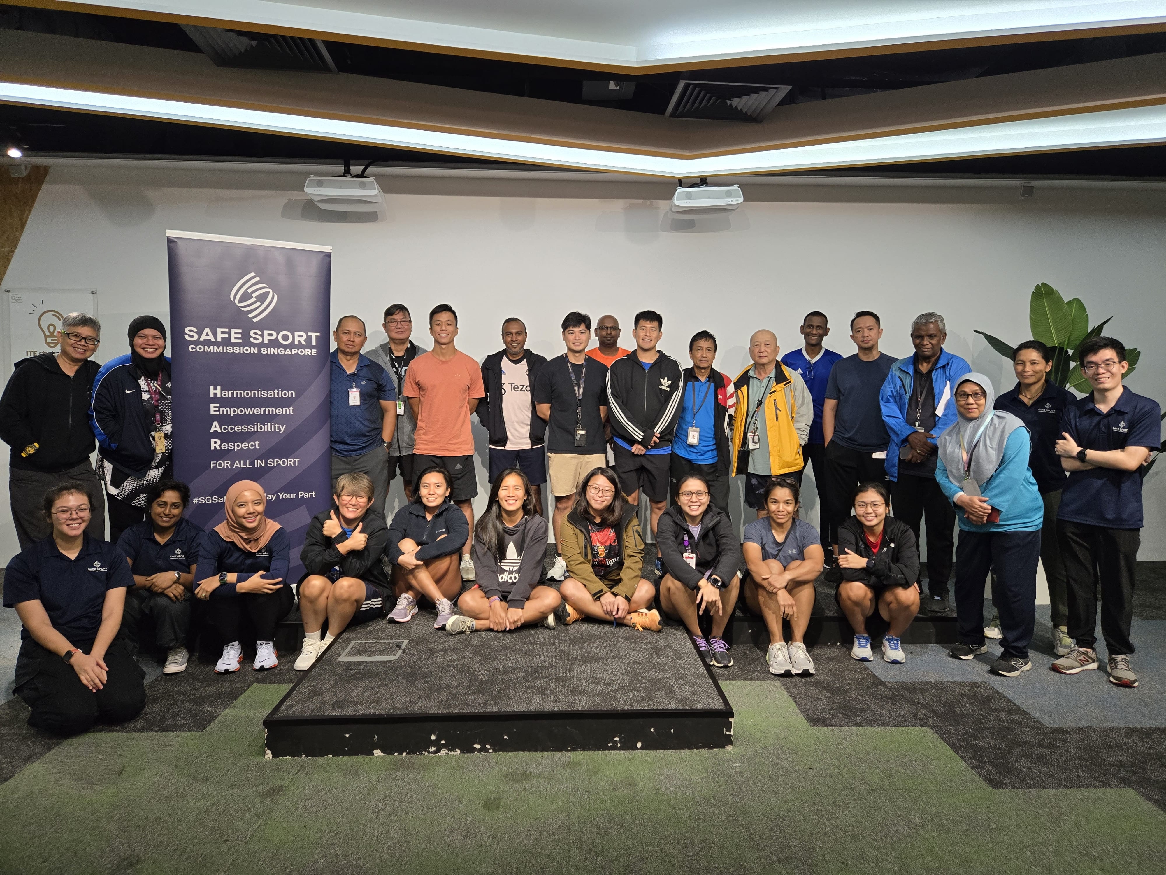 A group photo of 23 people, standing or sitting, with a banner on the left reading "Safe Sport Commission Singapore."