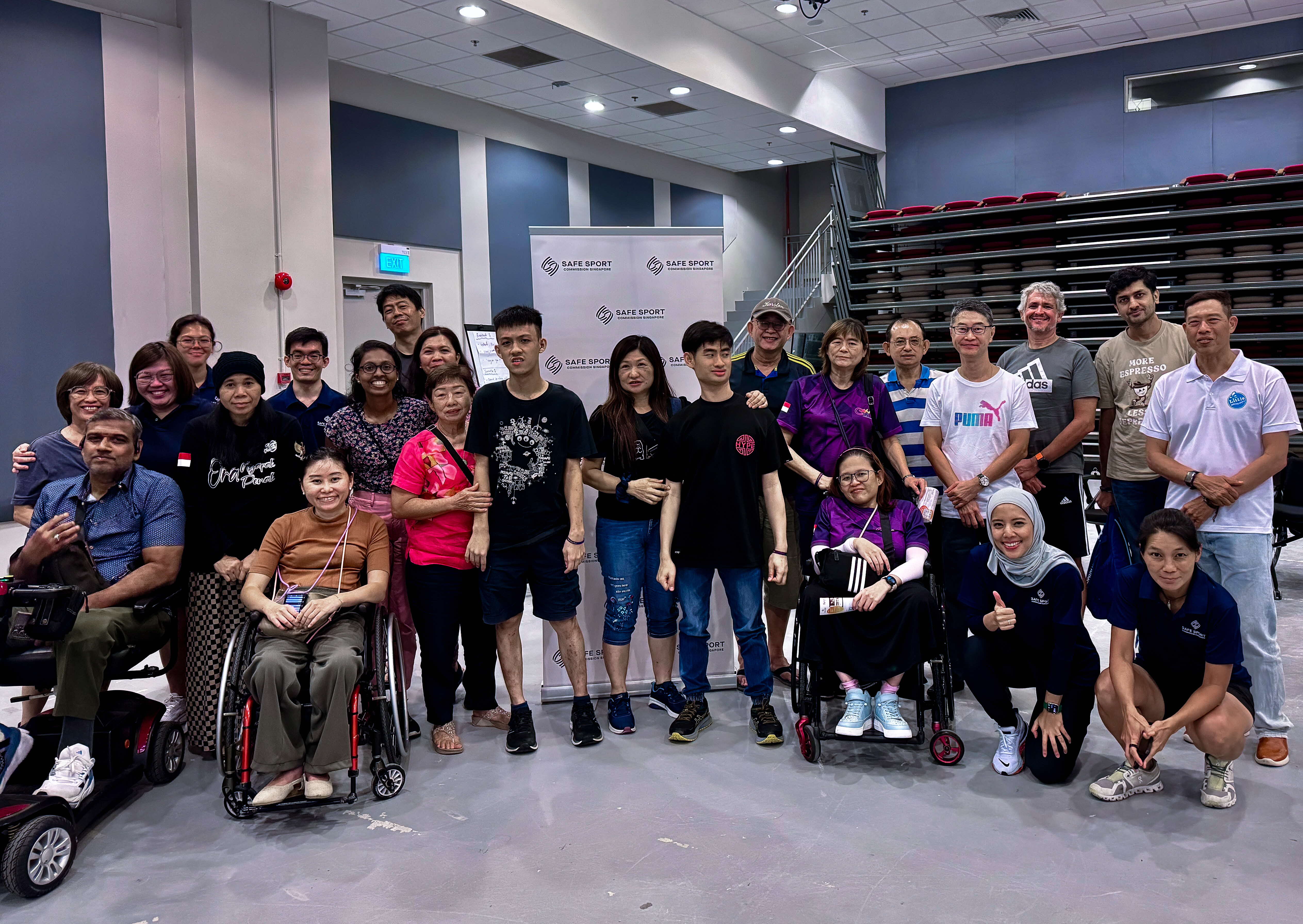 A group of people, some seated in wheelchairs, standing indoor in front of a Safe Sport banner, and near tiered seating.