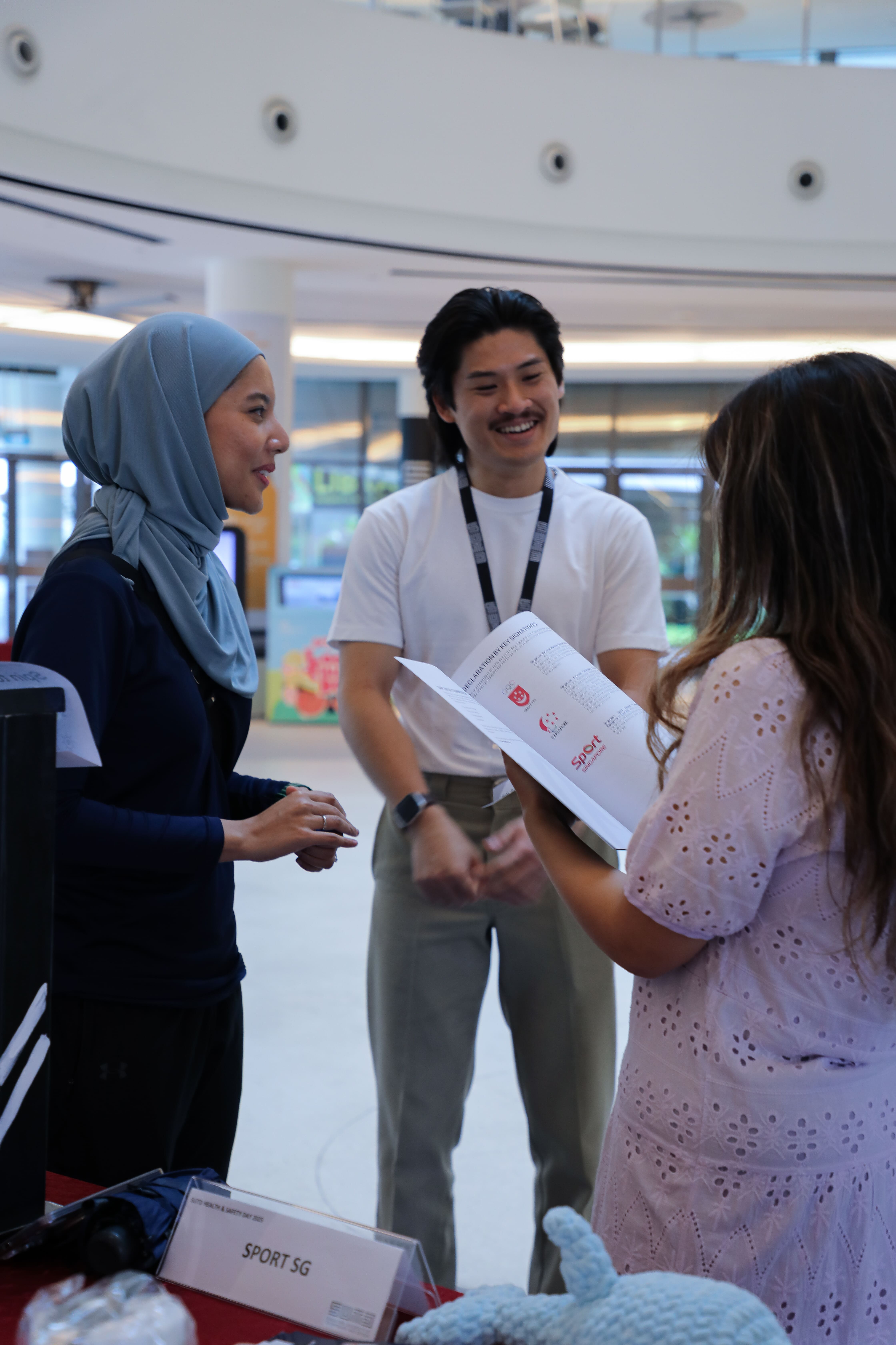 Three people are standing indoors, with one person wearing a headscarf and another holding a brochure from Sport SG.