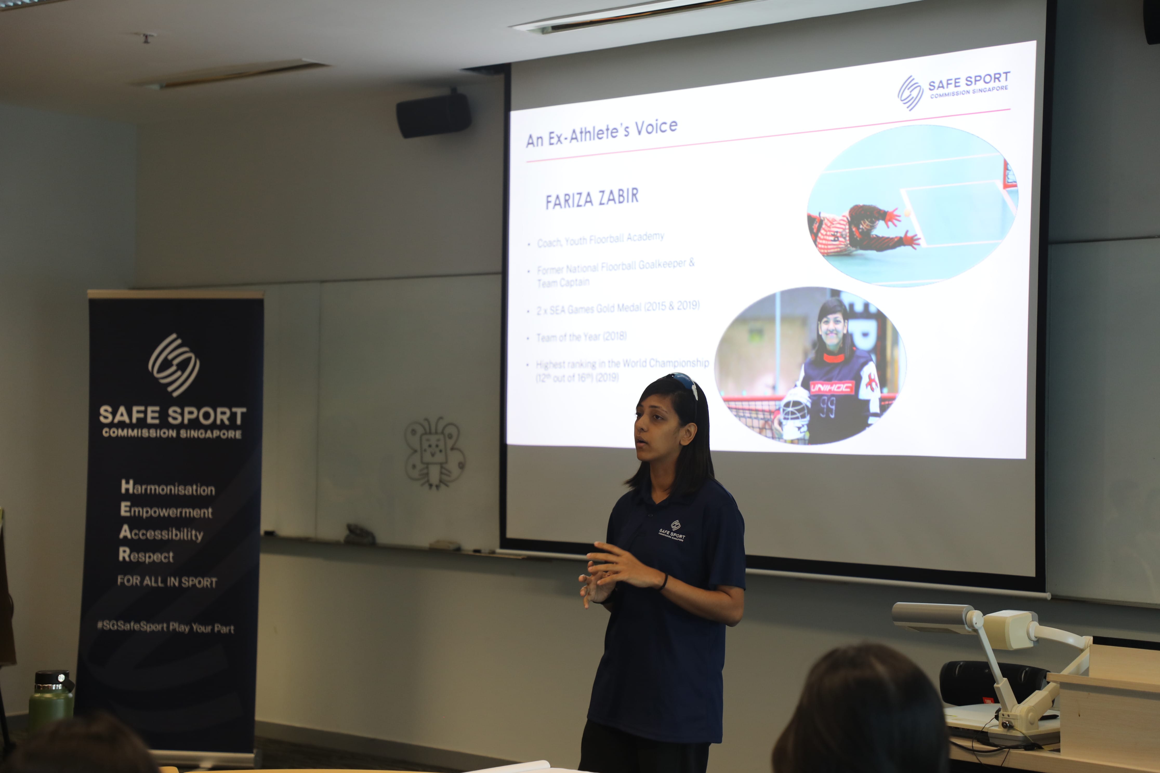 A person in a navy shirt stands in front of a projector screen and a banner with "SAFE SPORT COMMISSION SINGAPORE."