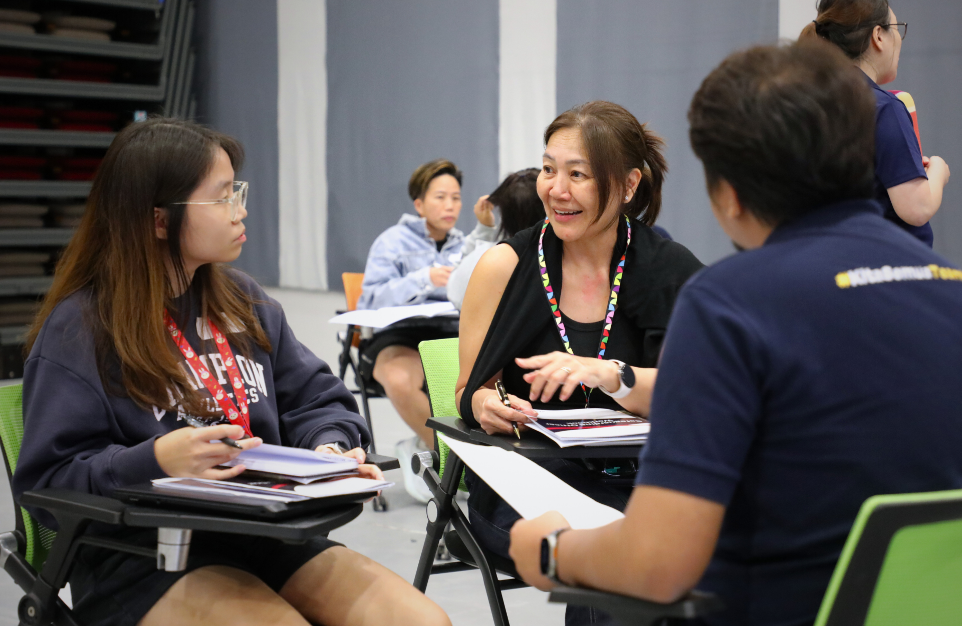 People in a classroom setting engaged in discussion. A woman in the center smiles while speaking. There are notebooks and pens, suggesting a collaborative atmosphere.