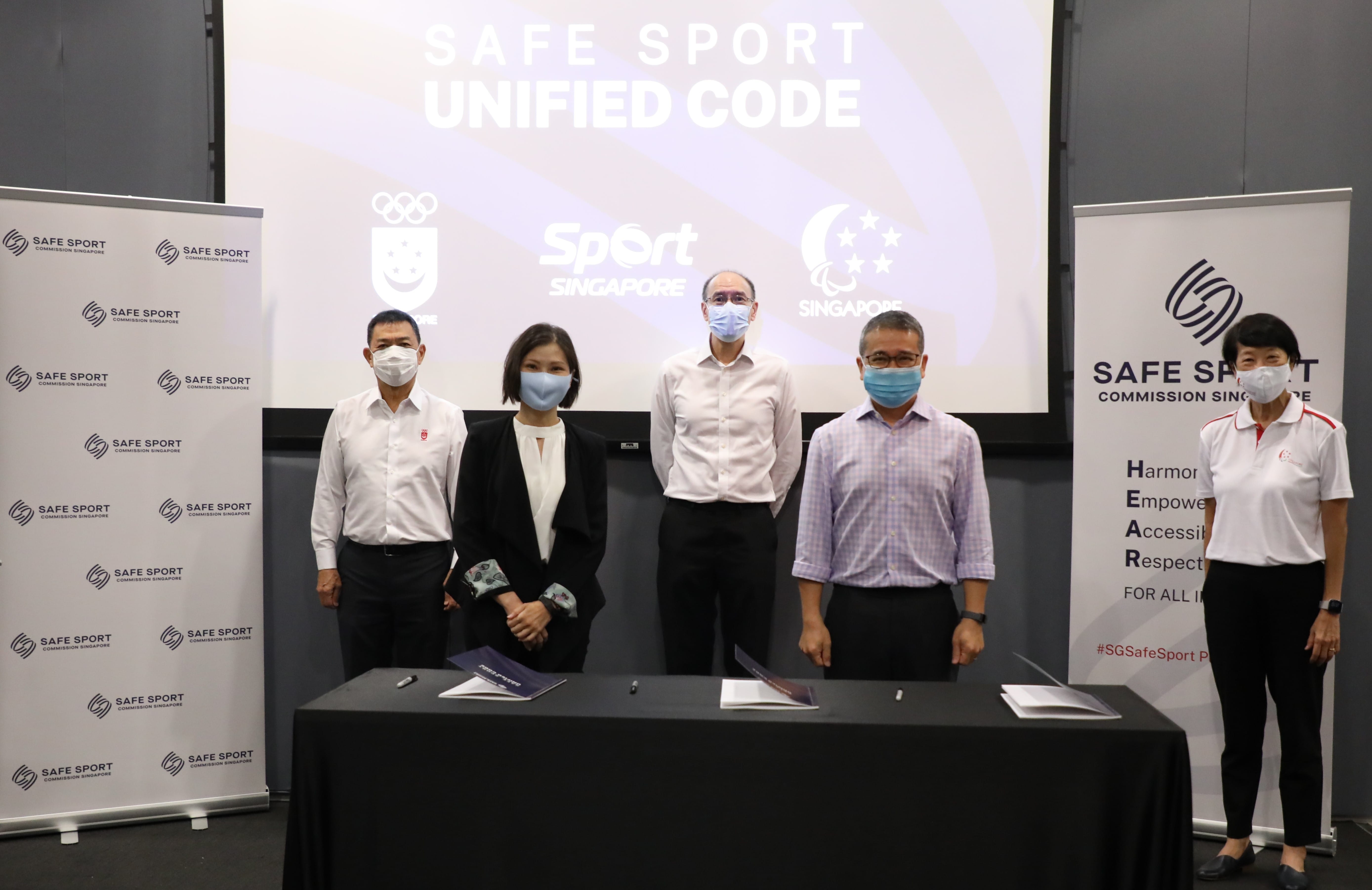 Five individuals stand behind a table, with the "SAFE SPORT UNIFIED CODE" logo displayed on the screen behind them.