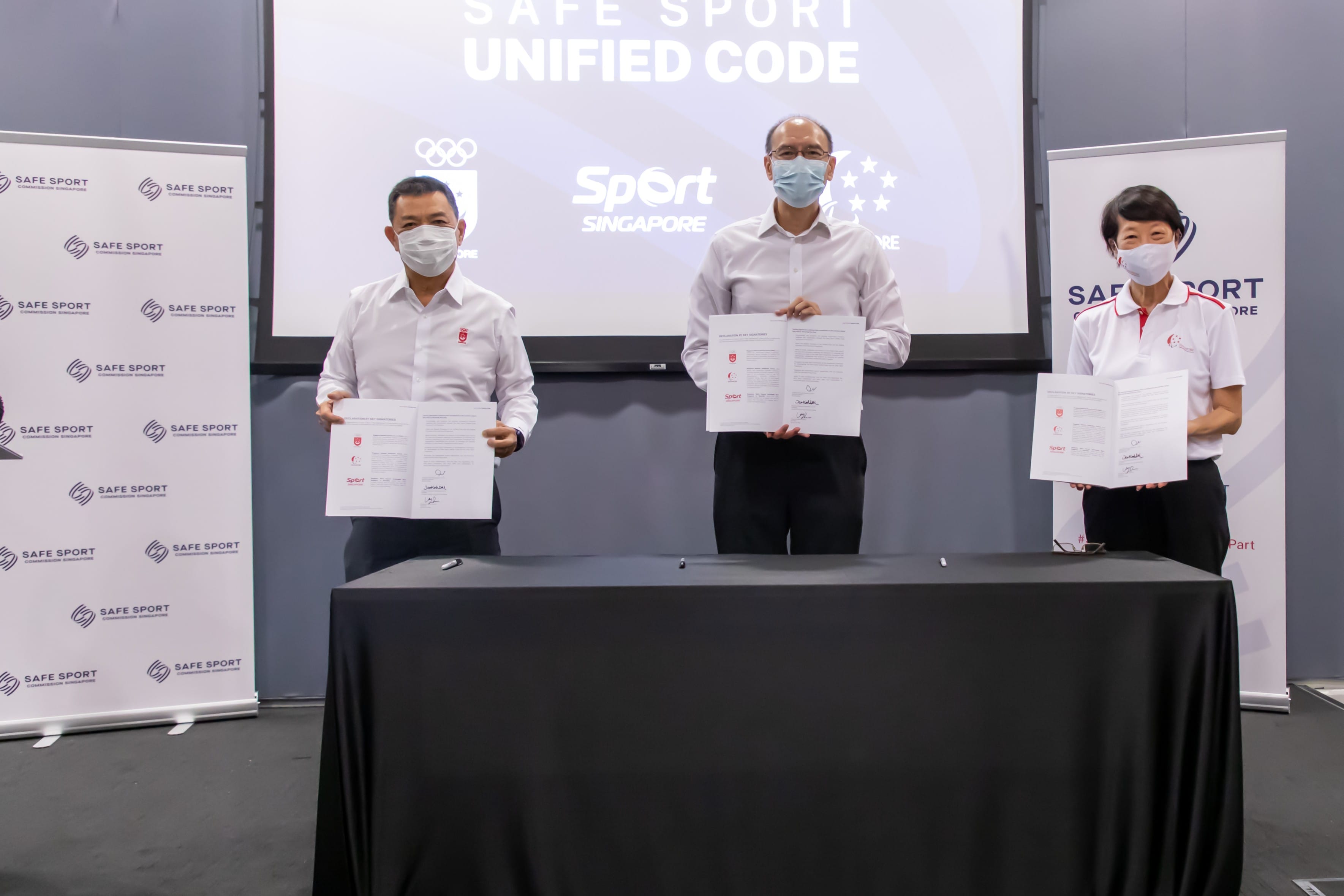 Three people in formal attire stand holding documents in front of a projection screen reading "Safe Sport Unified Code".