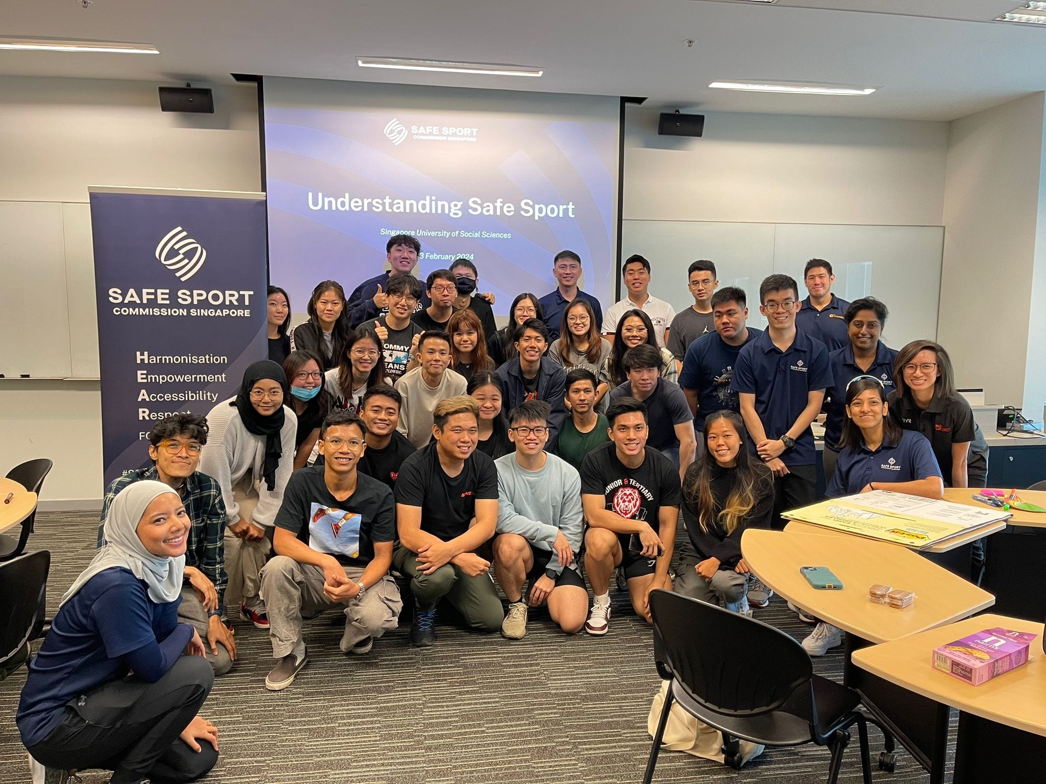 Group of people posing in a classroom, with a banner that reads "Safe Sport Commission Singapore" and a presentation screen.