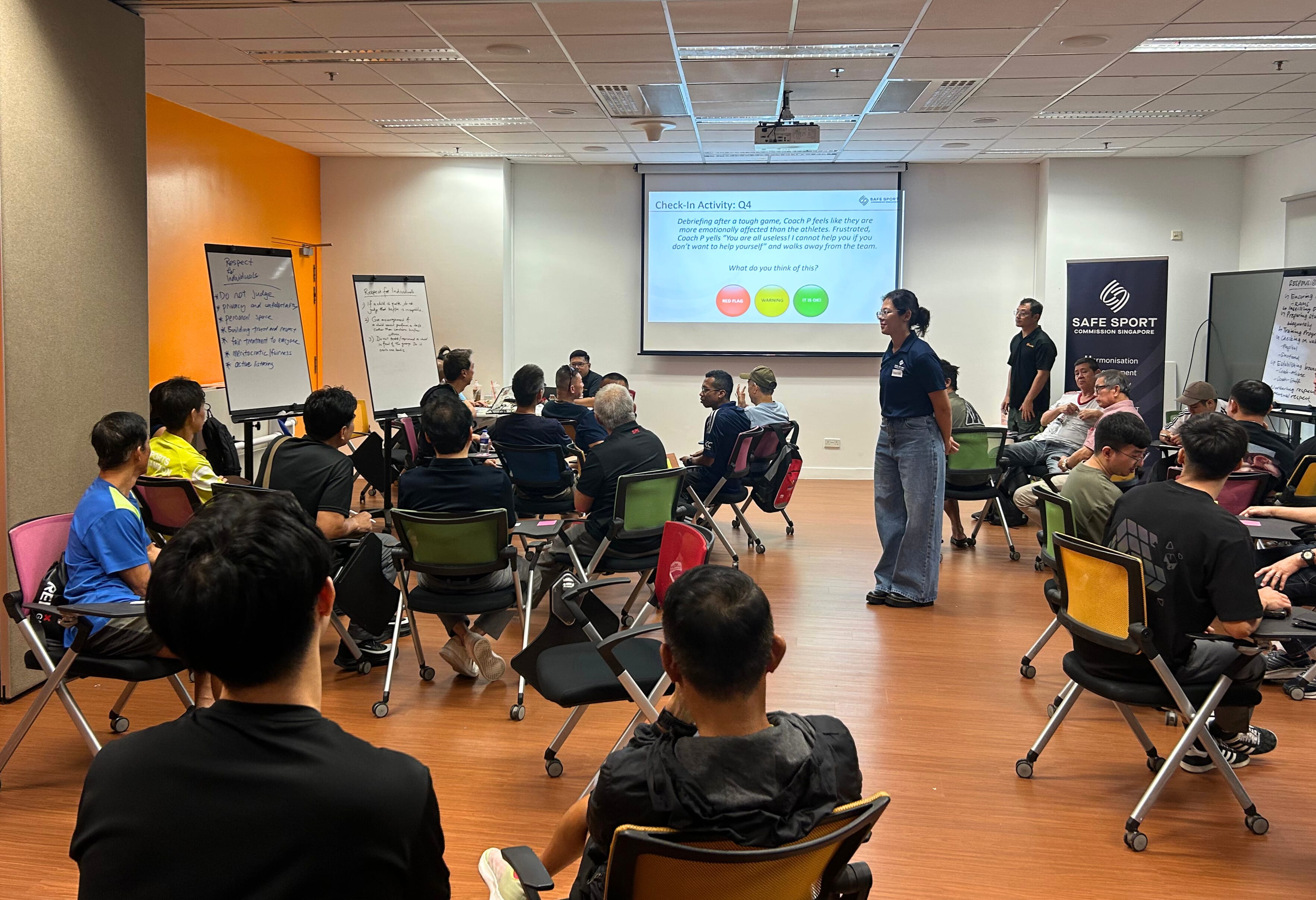 A group of people sitting in a classroom, facing a presentation screen and flip charts, with one person standing.
