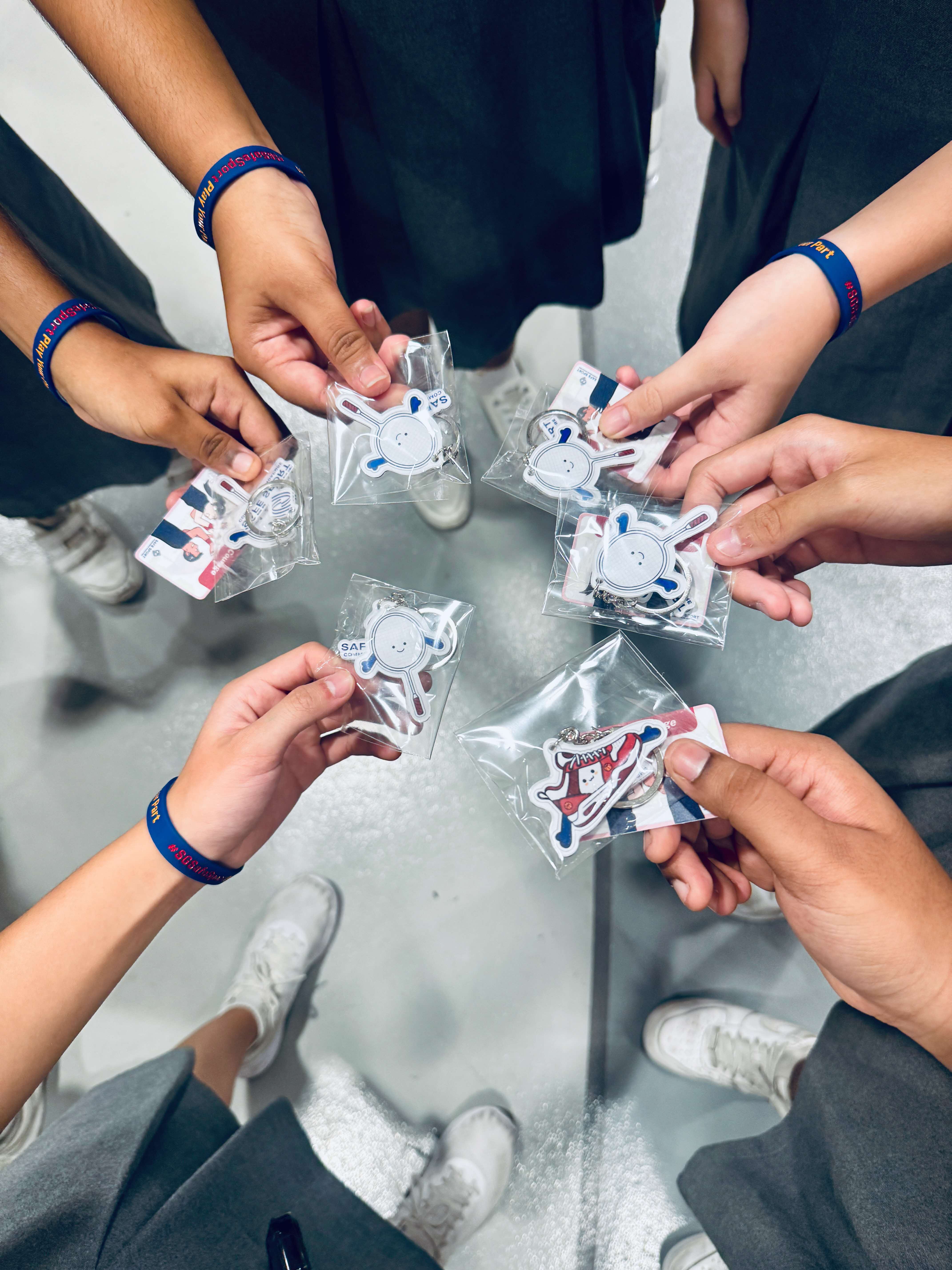 Six hands holding cute keychains form a circle. Each person wears a blue wristband and white sneakers, conveying a sense of unity and friendship.