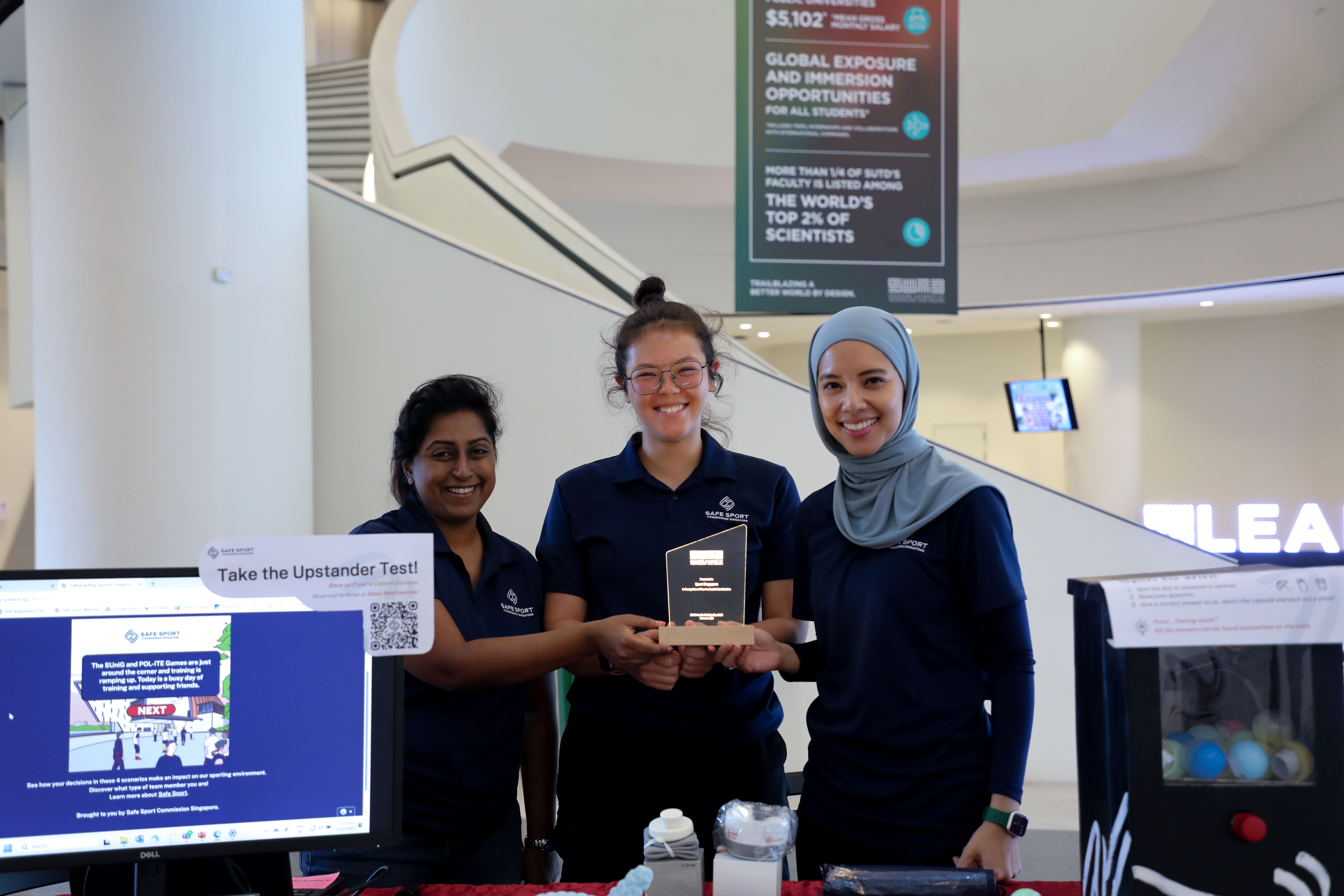 Three people in navy shirts hold a trophy behind a booth with a laptop screen showing "Take the Upstander Test!".