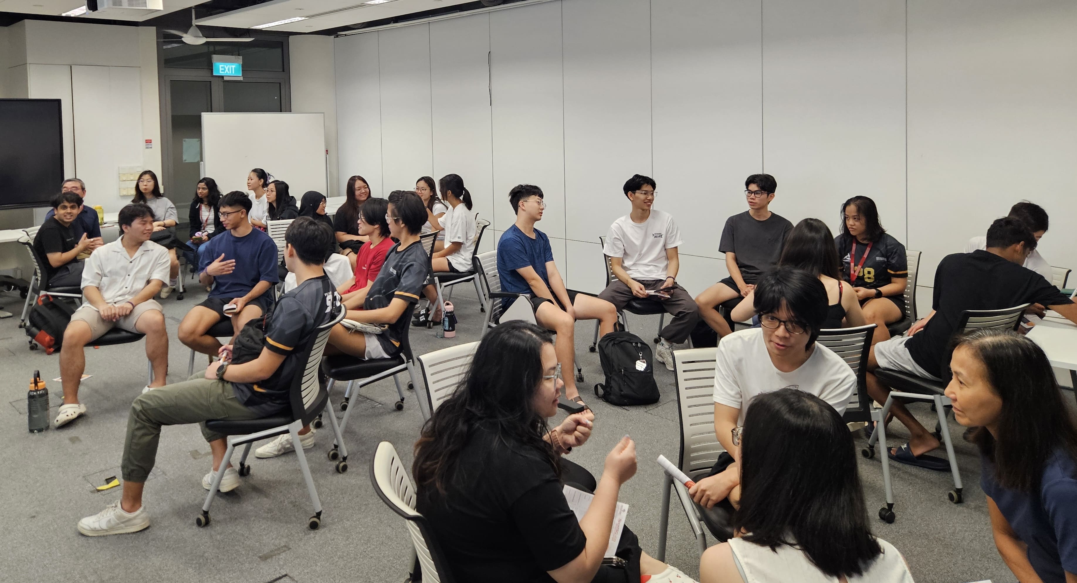 A group of young adults sits in chairs arranged in a circle in a brightly lit room with a whiteboard and television.