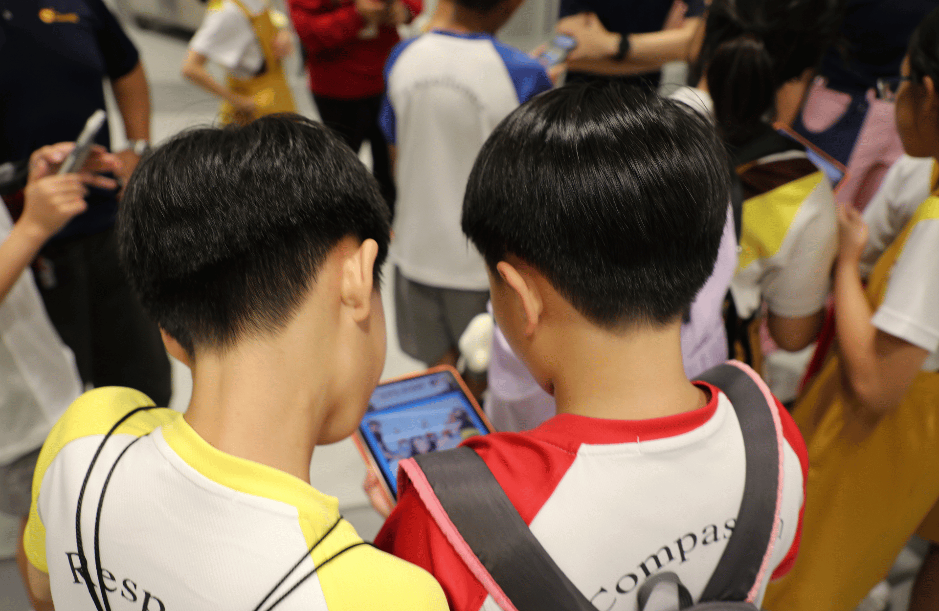 Two children wearing backpacks stand closely together, viewing a tablet screen. They are in a group setting, suggesting a lively, social environment.