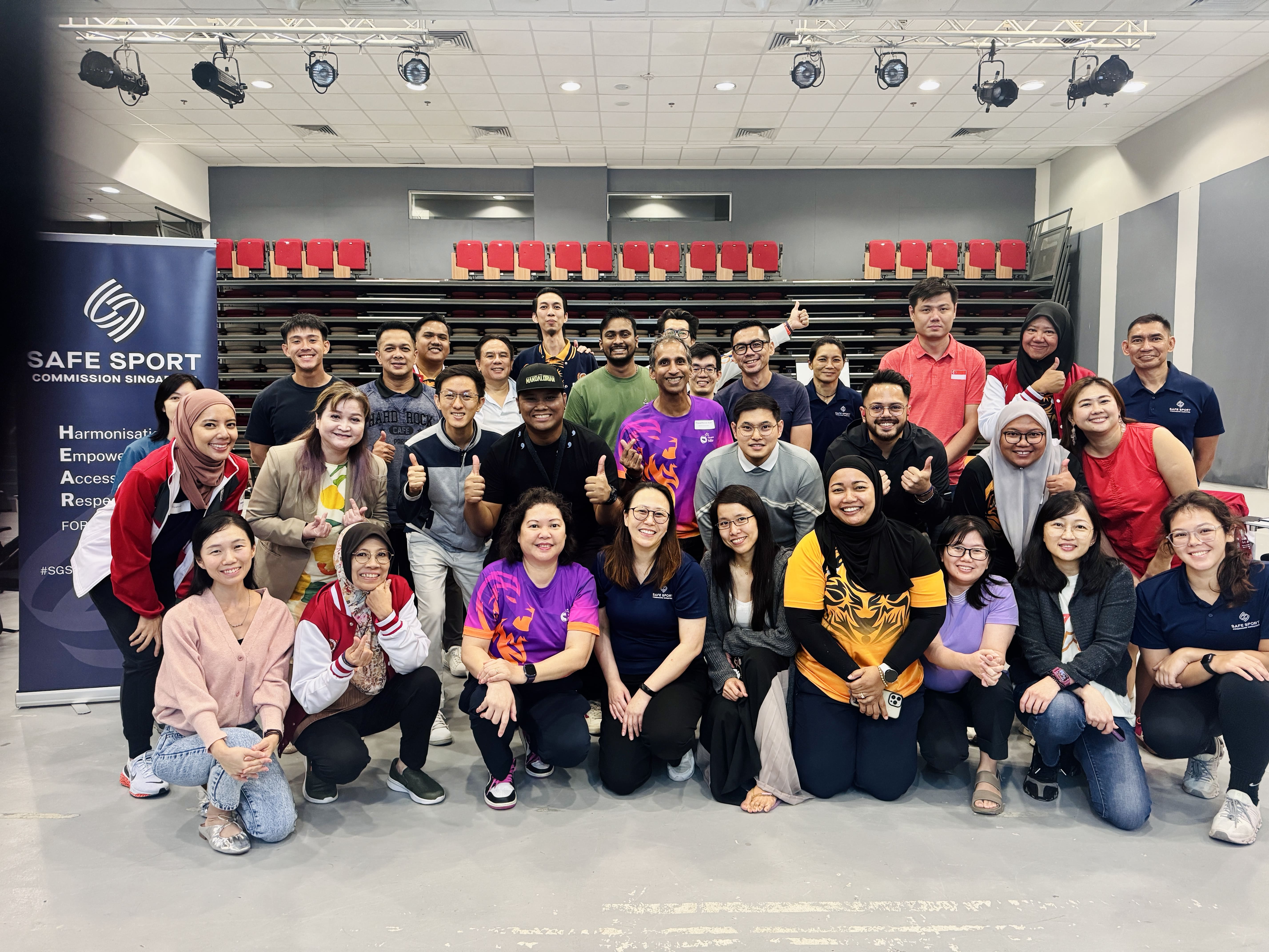 A large group of people in various clothing poses for a photo in a room with red seats and a Safe Sport banner.