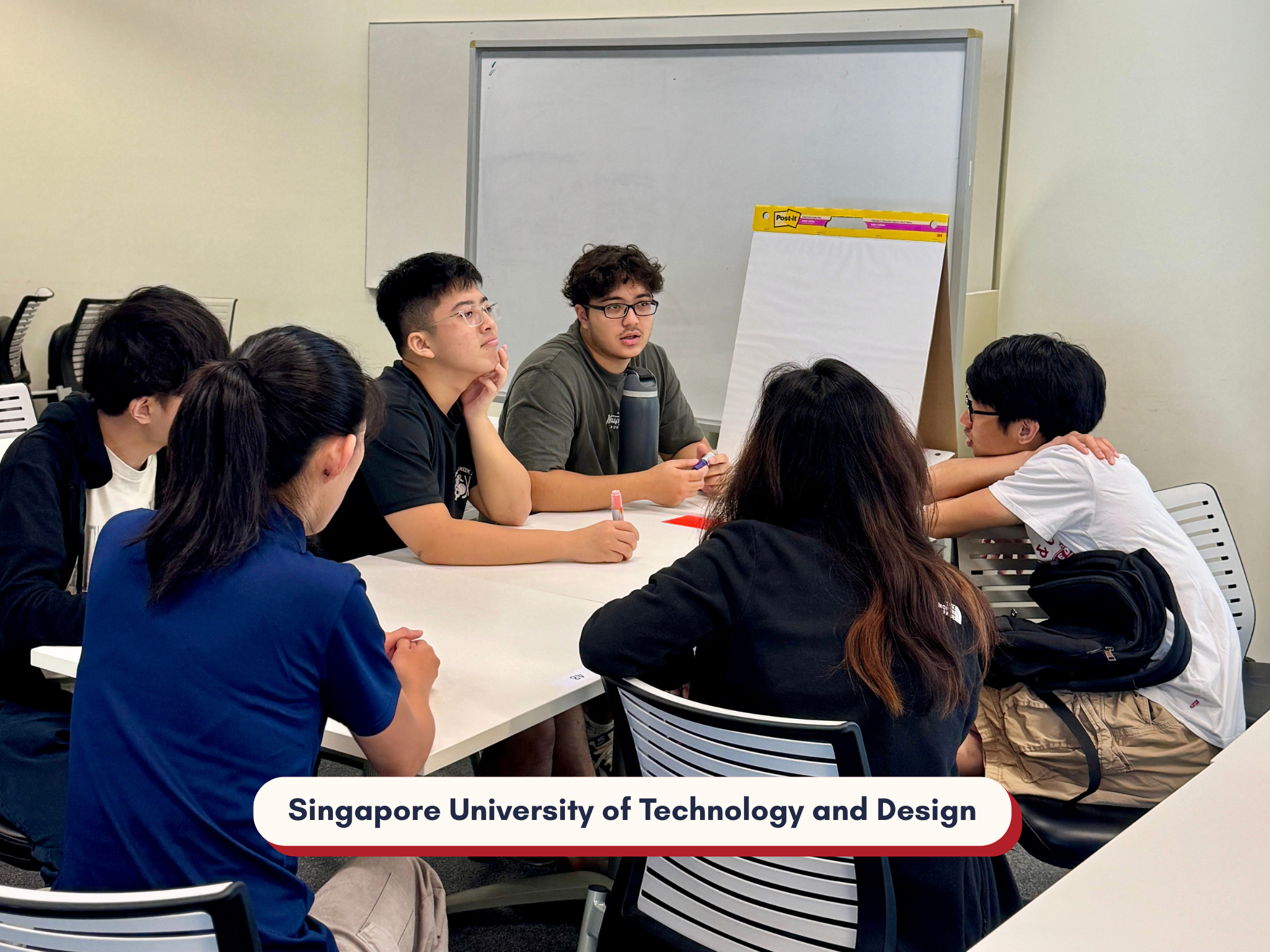 A group of six students sits around a table, engaged in discussion. A blank flipchart is nearby, suggesting a brainstorming session. The setting is the Singapore University of Technology and Design.