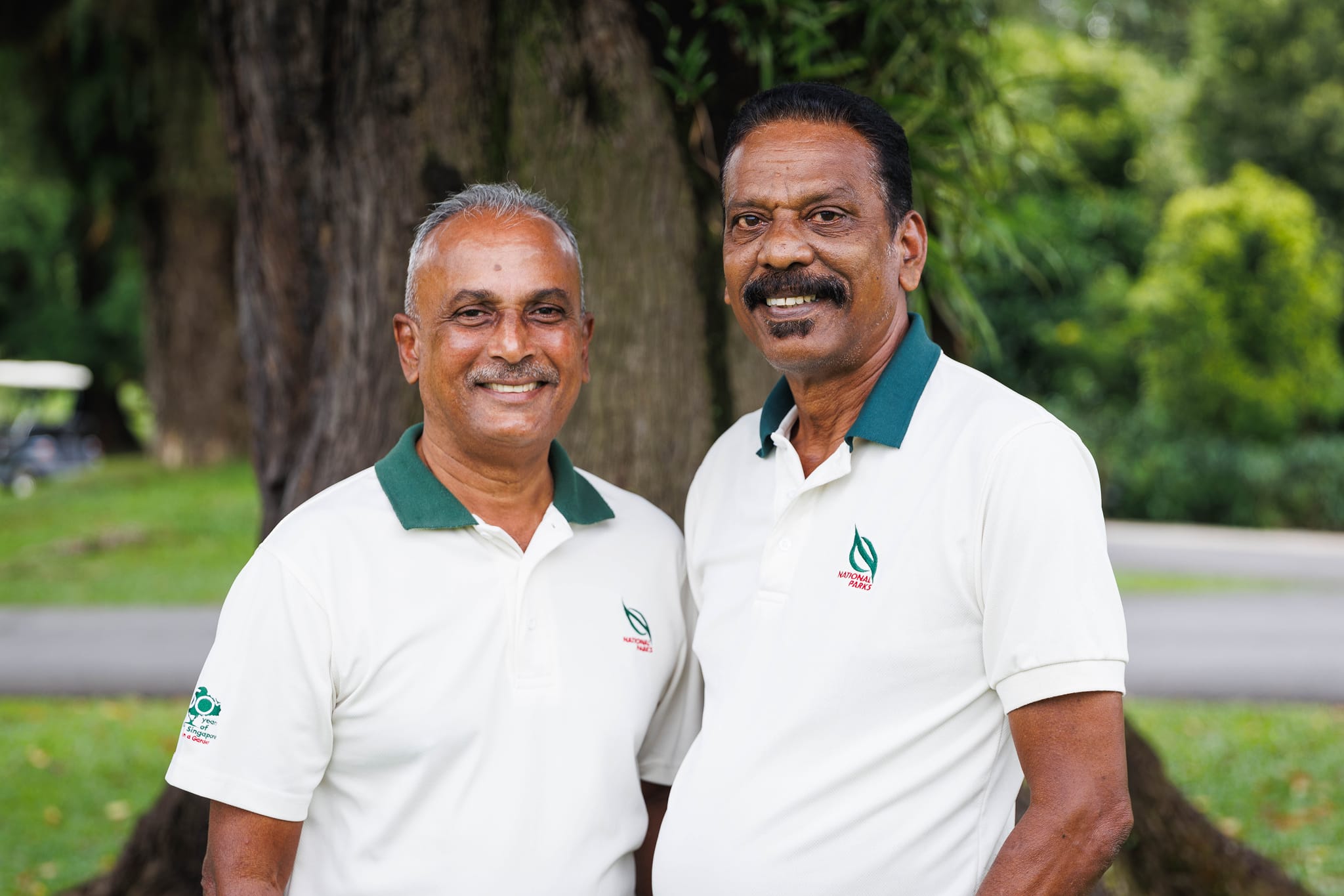 Two men wearing National Parks polo shirts stand outdoors in front of a tree.
