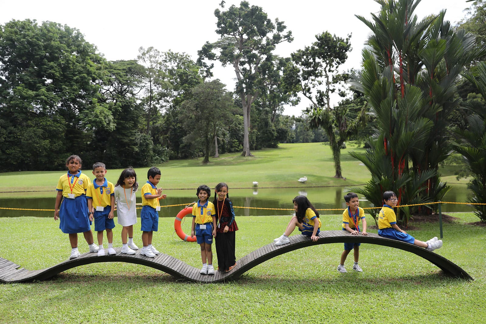 Children in yellow and blue school uniforms on a wooden bridge in a park. A pond and greenery in the background.