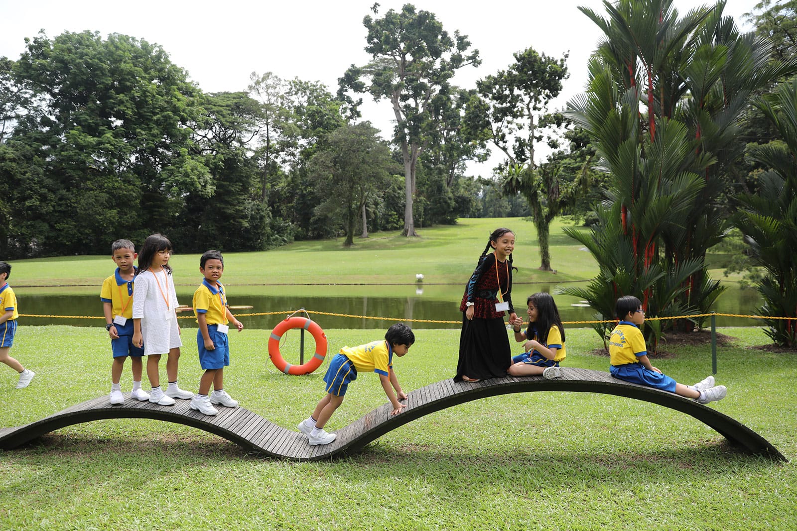 Children in school uniforms on a curved bridge in a park, pond and trees in background.