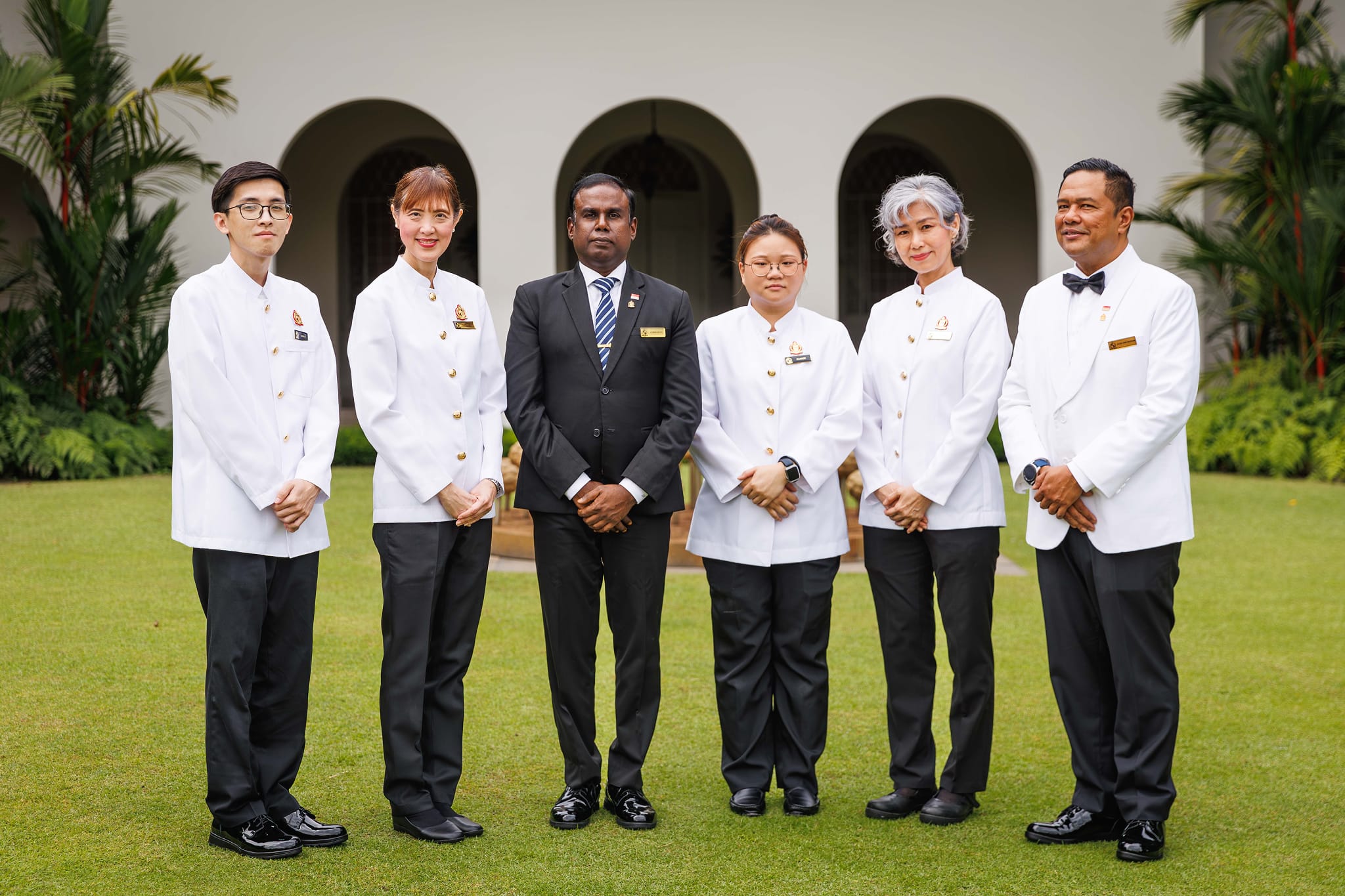 Six people in uniforms stand on grass in front of a building with archways.