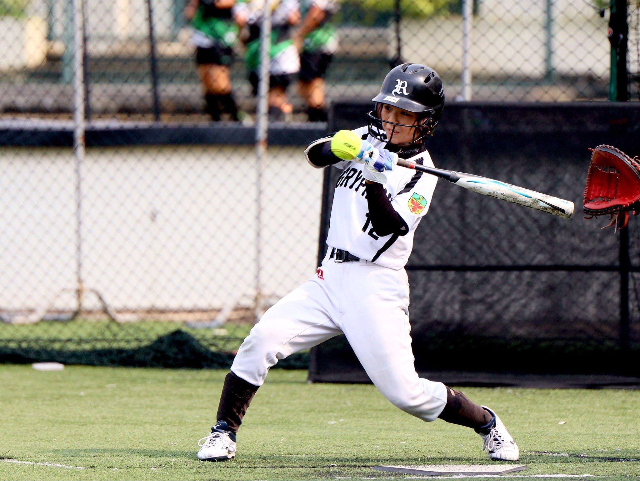A softball batter in white uniform and black helmet swings at a yellow ball near home plate on a green field.
