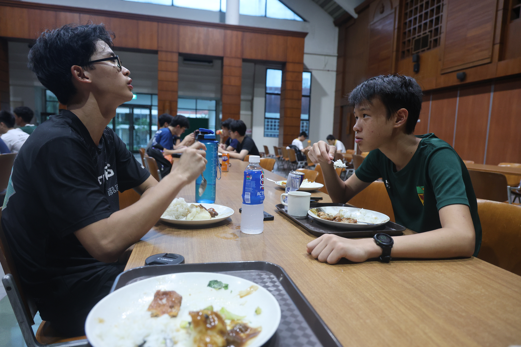 Two teenage boys eating rice meals on trays at a cafeteria table, talking to each other.