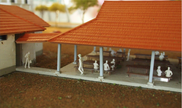 Miniature scale model of a covered outdoor area with red roofs, white figurines, and small benches.