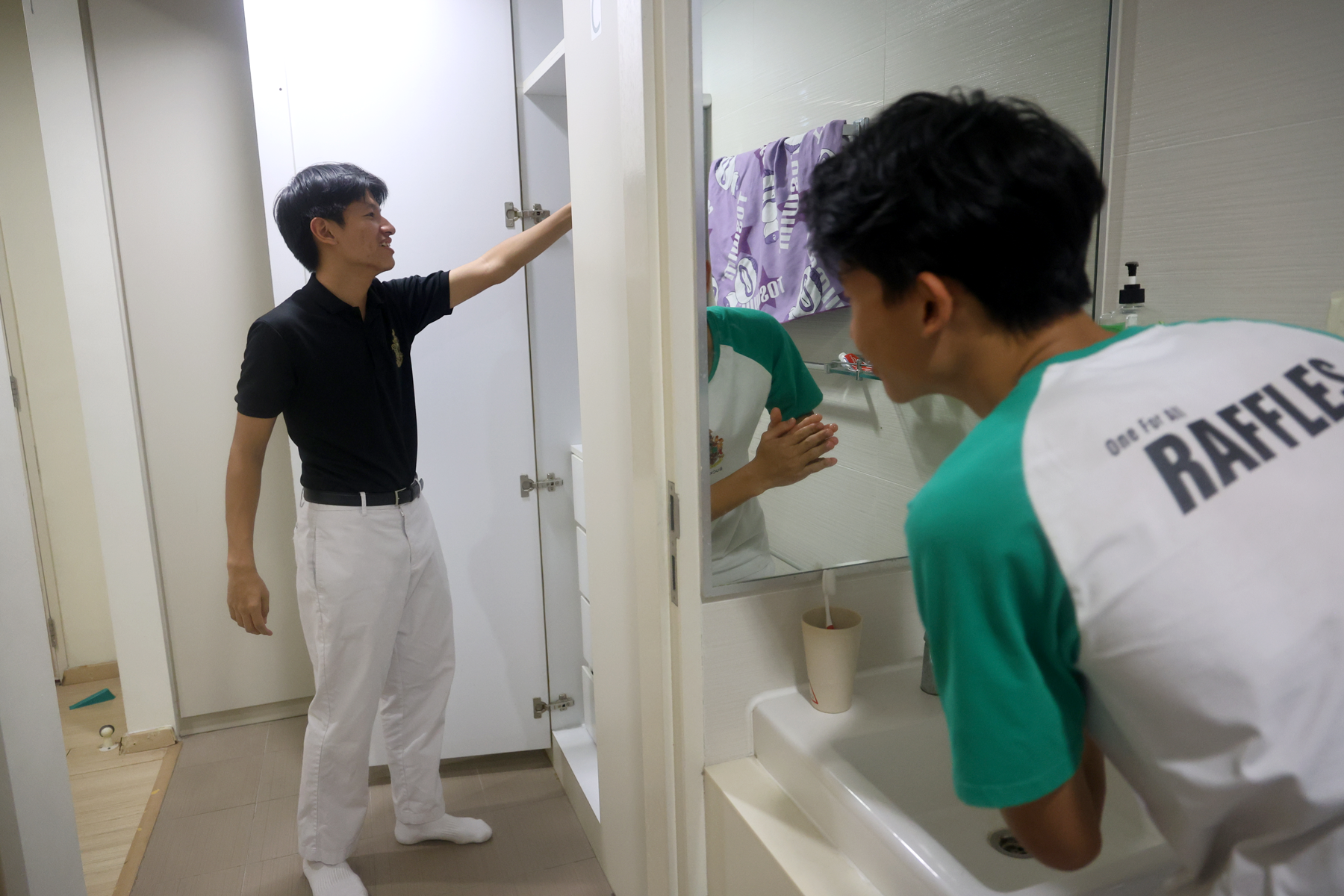 A man in black opens a cabinet while another in a Raffles shirt washes hands at a bathroom sink.