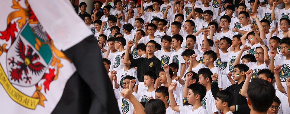 Large group of young students in white Helios shirts raising fists, with a black and white crest flag in foreground.