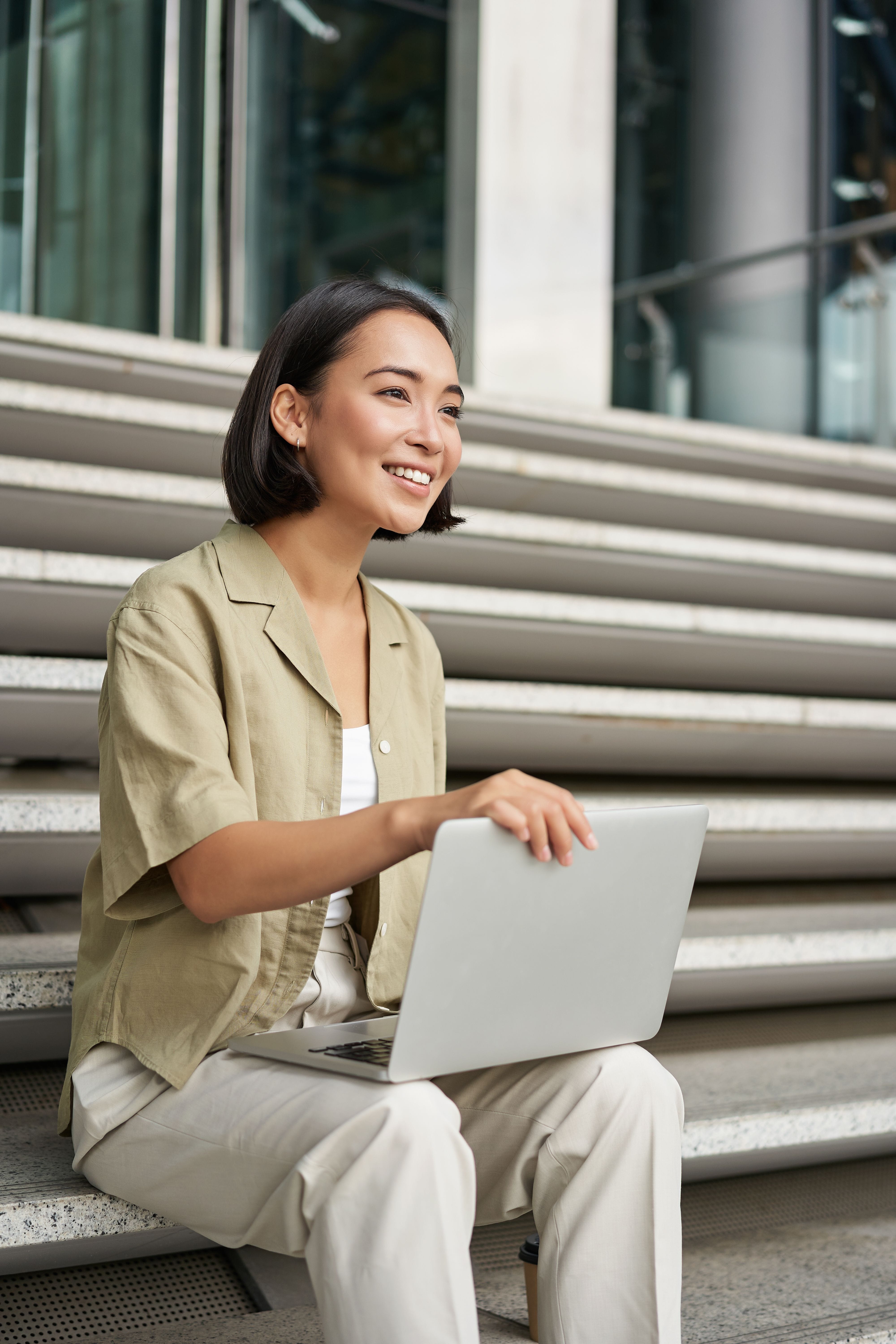 A smiling young woman with short dark hair sits on outdoor steps holding a laptop, wearing a green shirt.