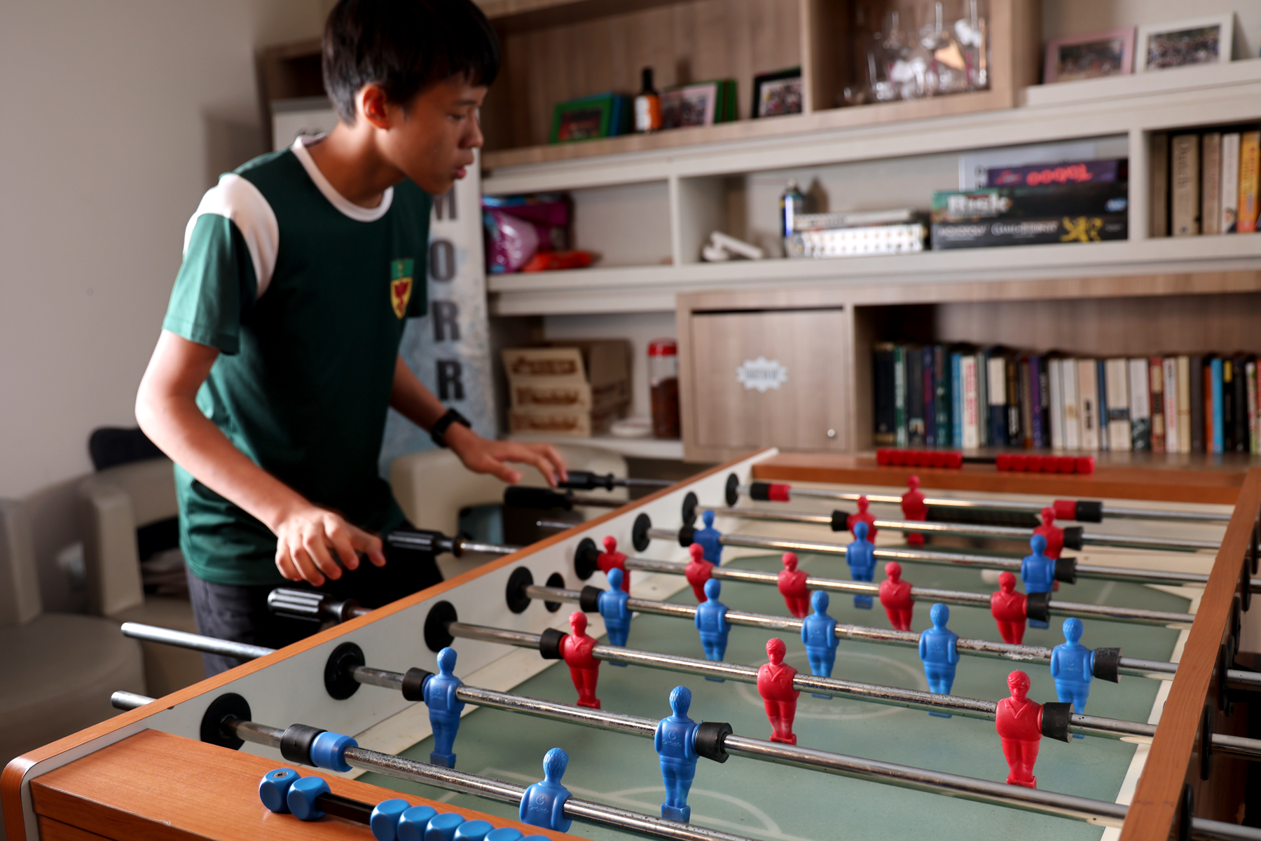 A boy in a green shirt plays foosball, gripping the rods over a table with red and blue player figures.