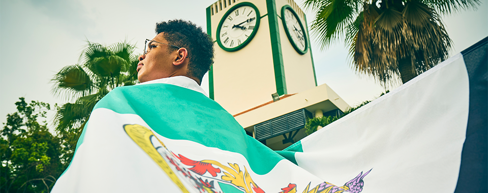 Young person draped in a Mexican flag stands before a clock tower surrounded by palm trees.