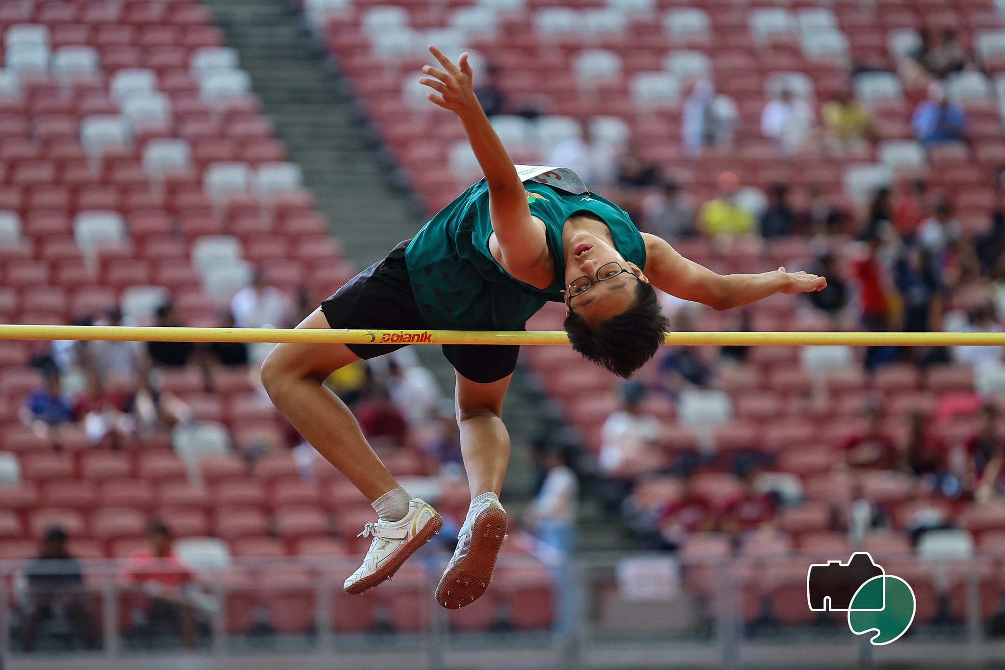A young male athlete wearing glasses clears a yellow high jump bar using the Fosbury flop technique in a stadium.
