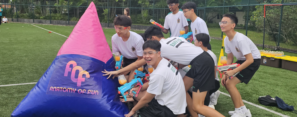 Teens in white shirts hold water guns behind a pink and blue inflatable barrier on a green field.
