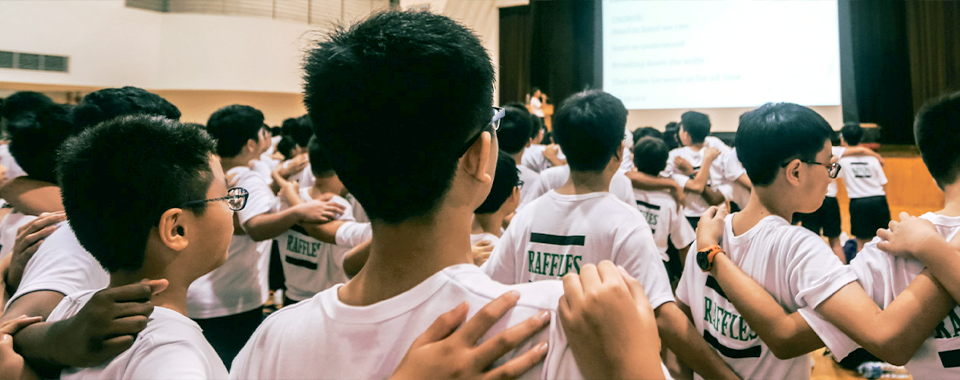 Students in white Raffles shirts stand in rows with hands on each other's shoulders in a school hall.