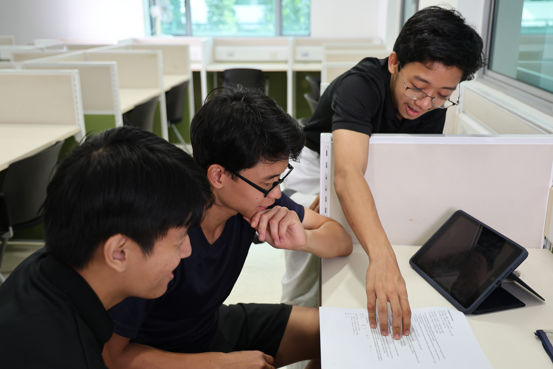 Three young men huddle over a printed document and tablet at a study desk in a bright room with cubicle dividers.