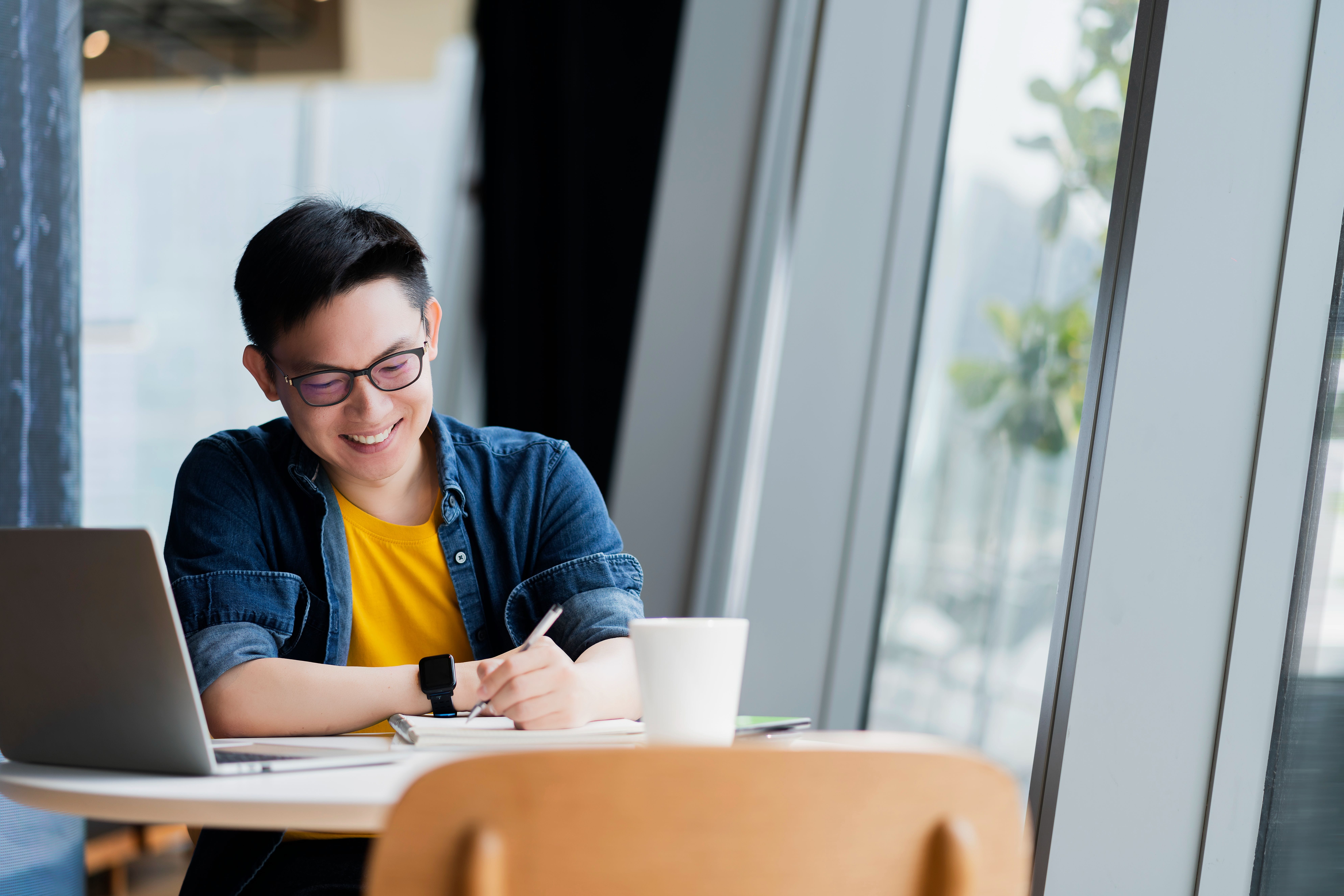 Smiling young man with glasses writing notes at a table with a laptop and white mug near large windows.