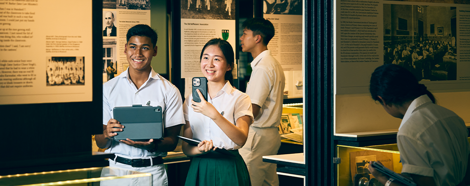 Four students in school uniforms explore a museum exhibit, two smiling in front holding a tablet and phone.