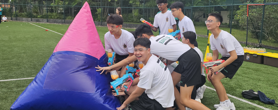Teens in white shirts hold water guns behind a pink and blue inflatable barrier on a green field.
