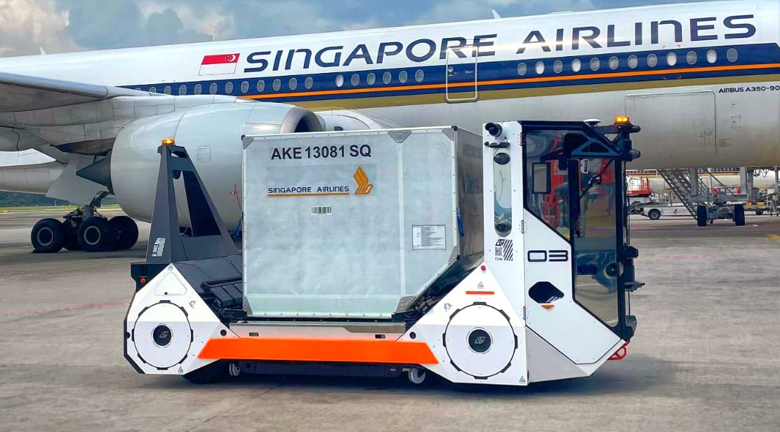 Self-driving baggage tractor towing a Singapore Airlines container near a parked aircraft on the tarmac.