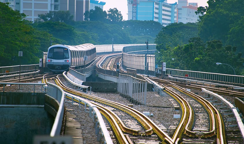 View of MRT tracks curving through greenery with a train moving toward residential buildings.